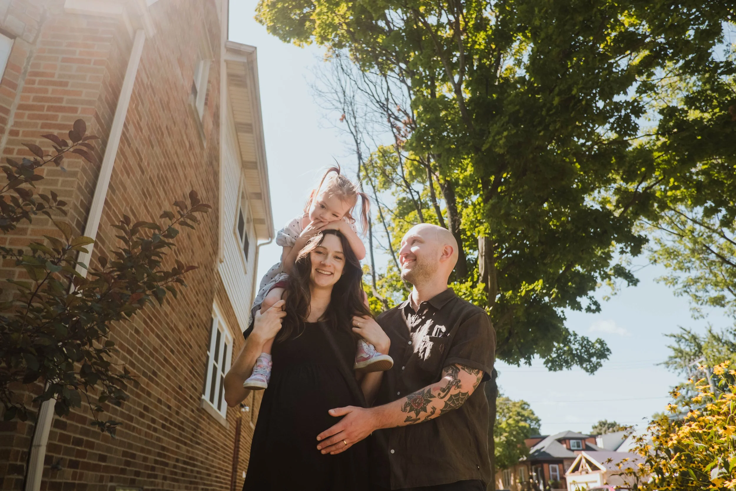 A family of three standing outdoors, with the mother carrying their young daughter on her shoulders, all smiling and enjoying a sunny day with green trees and a brick house in the background.