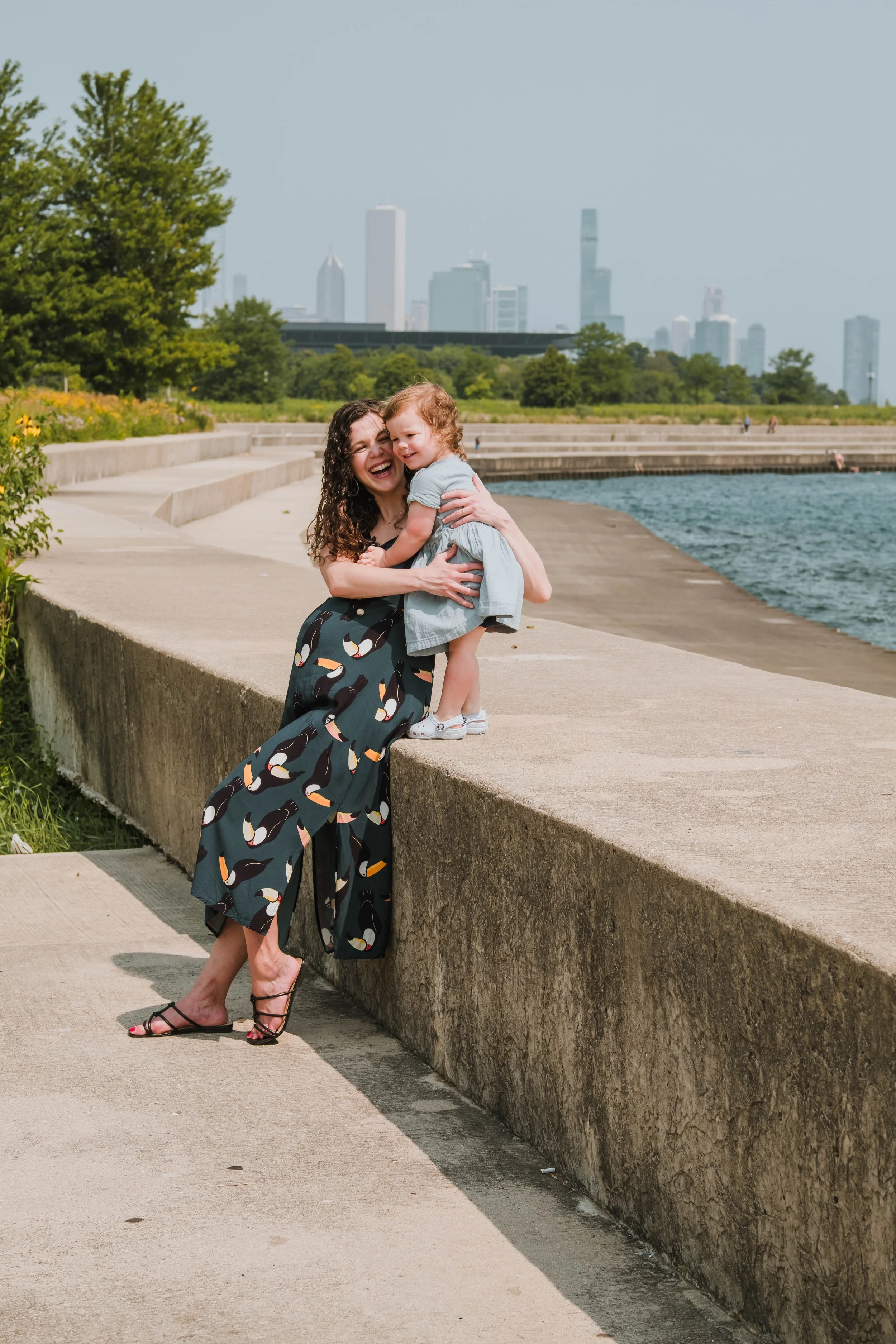 A woman in a zebra-patterned dress and black sandals is hugging a young girl in a light blue dress near a water body with city skyline in the background. The woman is smiling and the girl is happy.