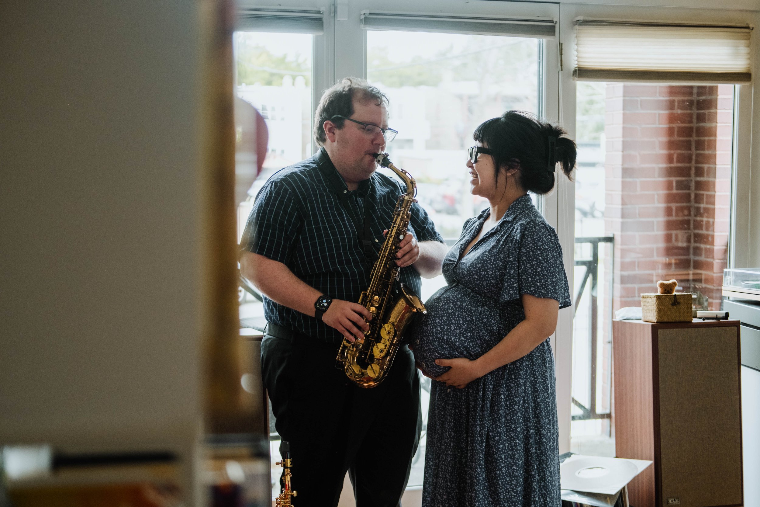 A man and a pregnant woman stand close together inside near large windows. The man is playing a saxophone, and the woman is smiling, with her hand resting on her baby bump. The scene is warmly lit and cozy.