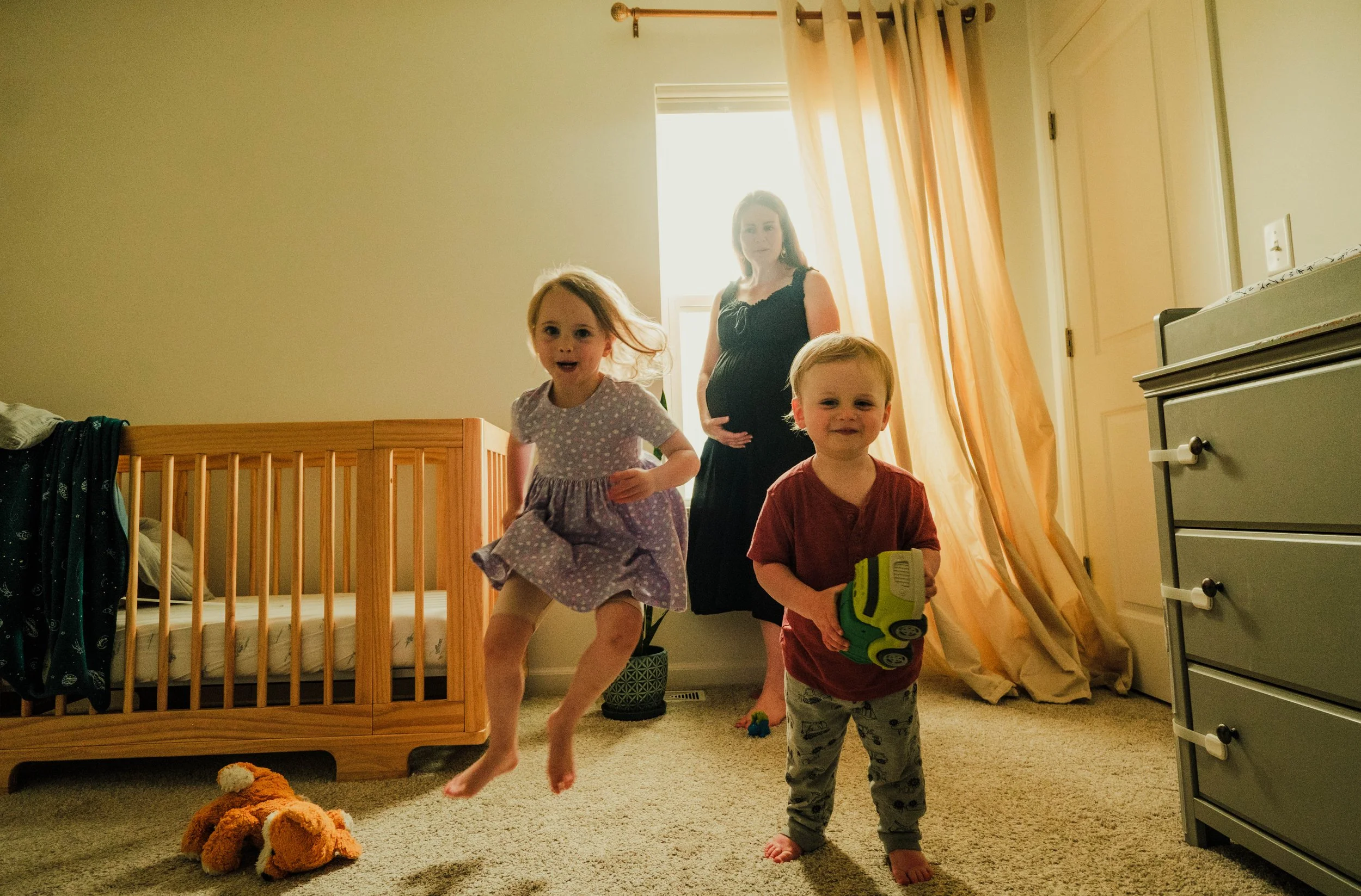 A pregnant woman and three children in a bedroom with a carpeted floor. One girl is mid-air jumping, and two boys are standing, one holding a green toy. The bed with a wood frame and a stuffed animal on the floor are visible, and sunlight filters thr