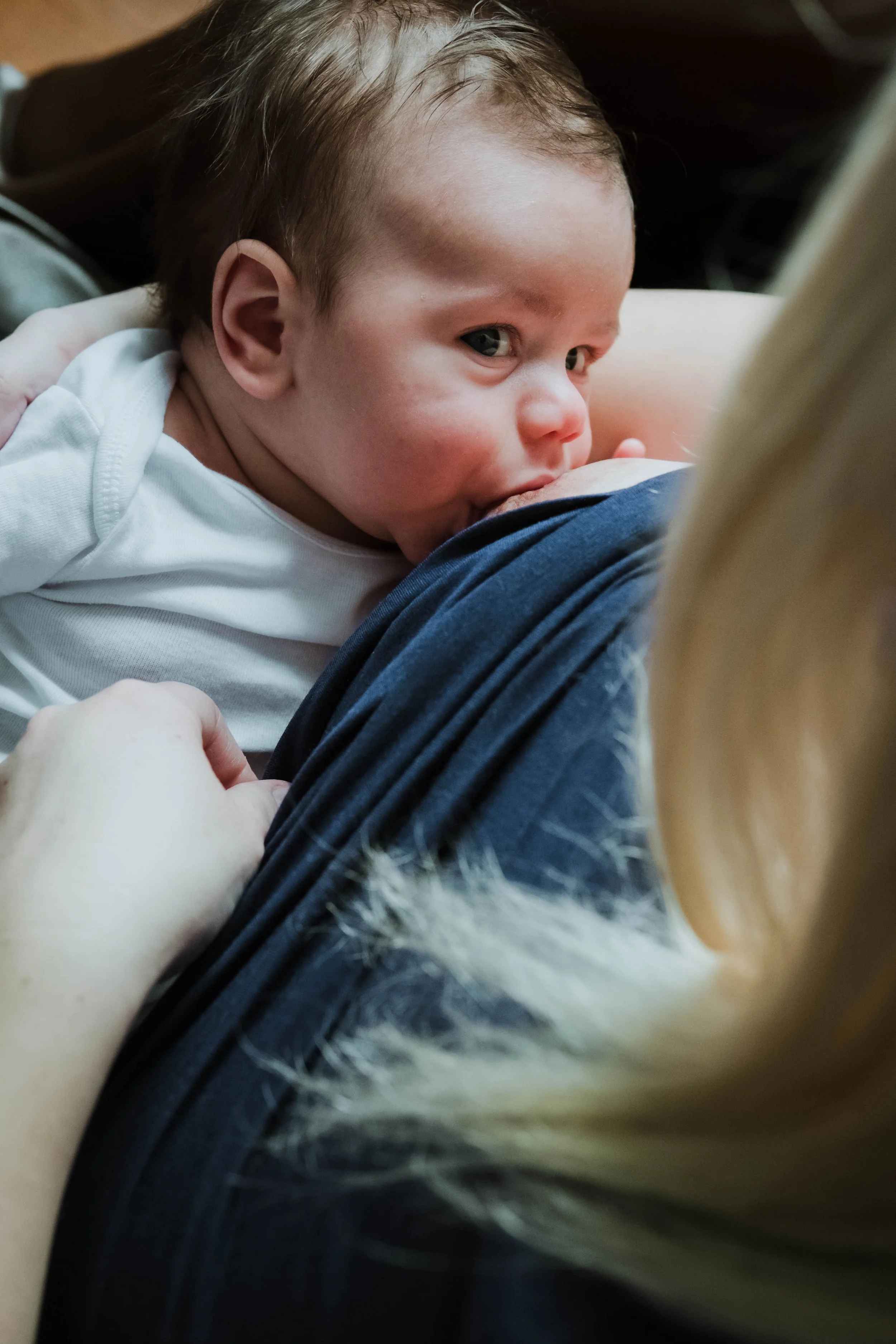 A young child with light brown hair, lying on an adult's lap, breastfeeding and looking at the camera with a gentle expression.