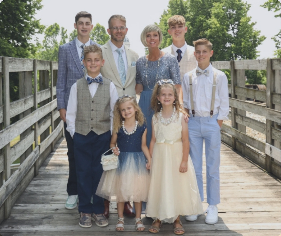 Family of nine posing on a wooden bridge outdoors, dressed in semi-formal attire for a special occasion.