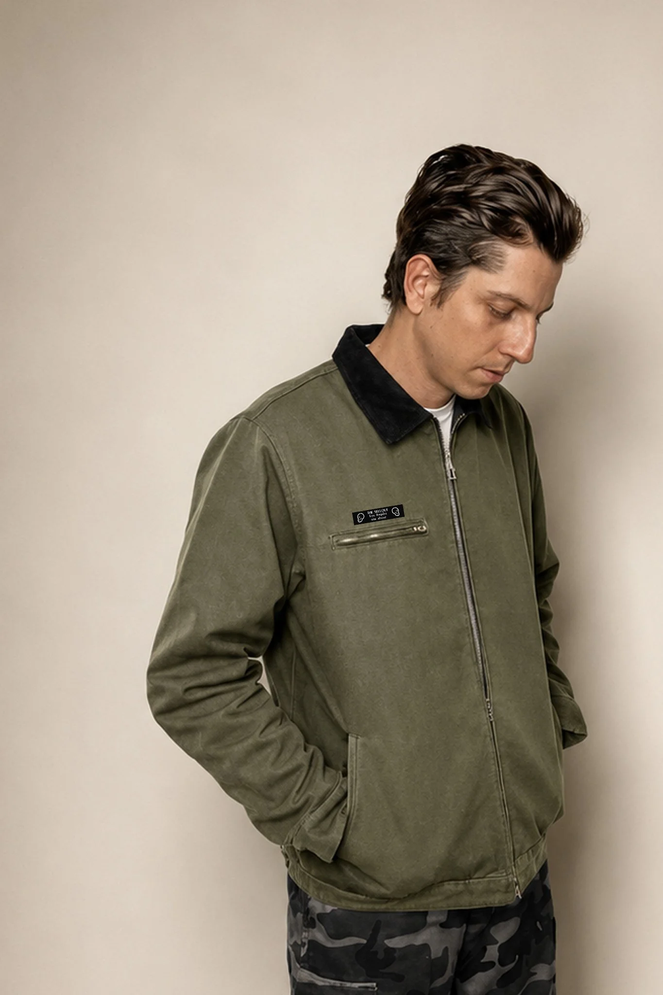 Model wearing The SellOut green canvas collar jacket with black corduroy collar, looking down in a minimalist studio portrait.