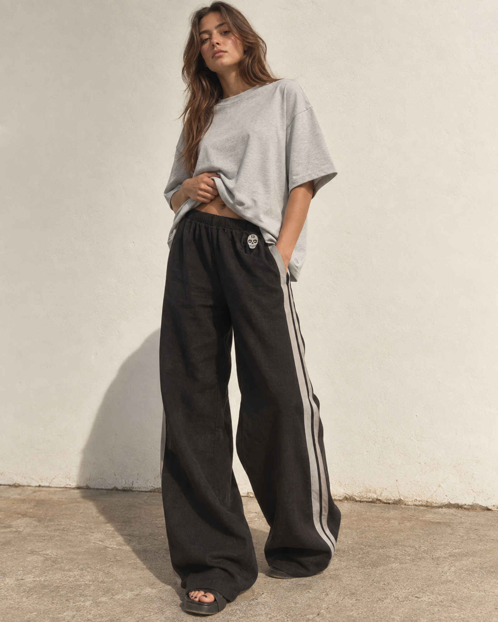 Woman wearing The SellOut black cotton-linen wide-leg track pants with double light-gray side tape and 13 Skull embroidery outdoors.