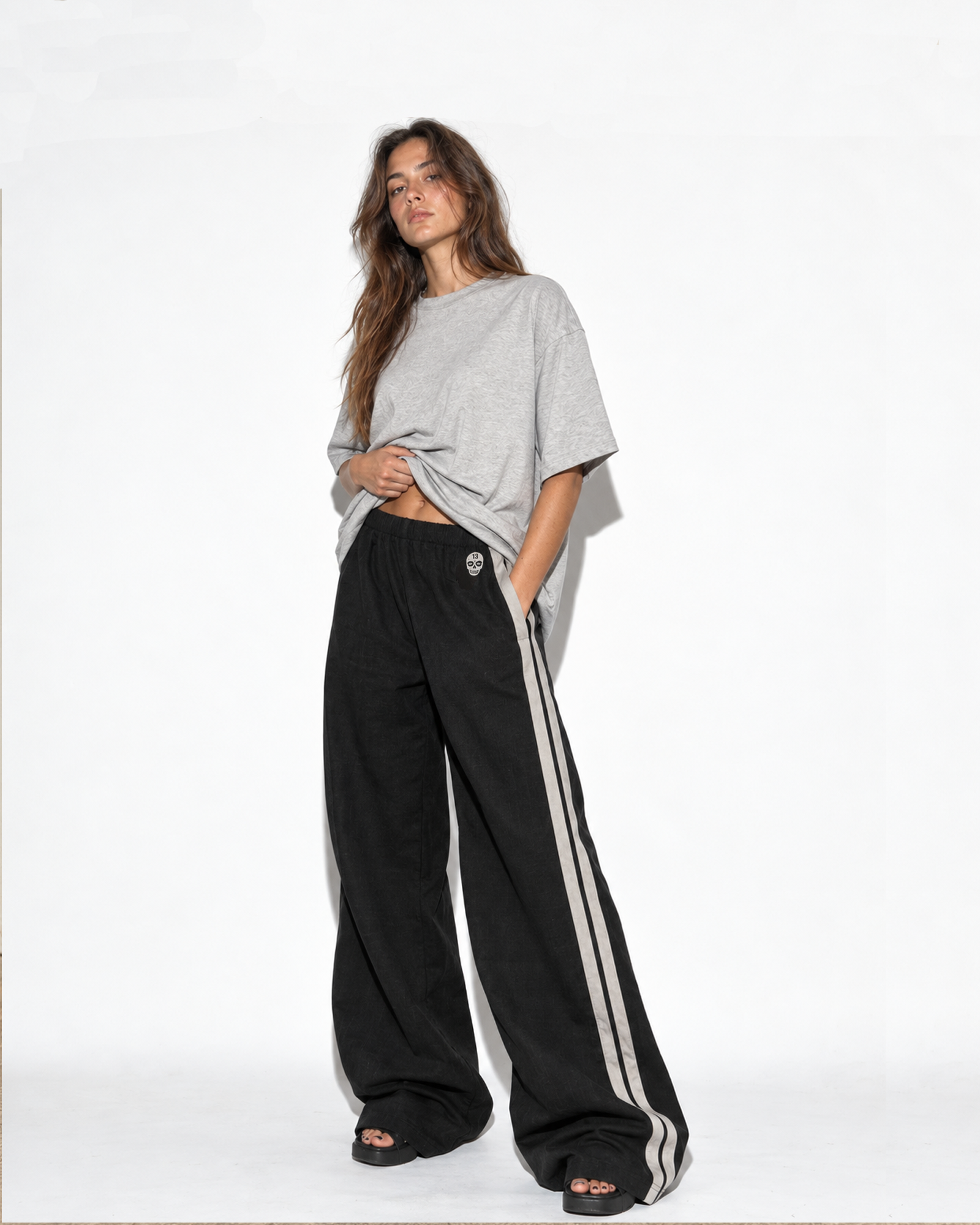 Woman wearing The SellOut black cotton-linen wide-leg track pants with double light-gray side tape and 13 Skull embroidery.