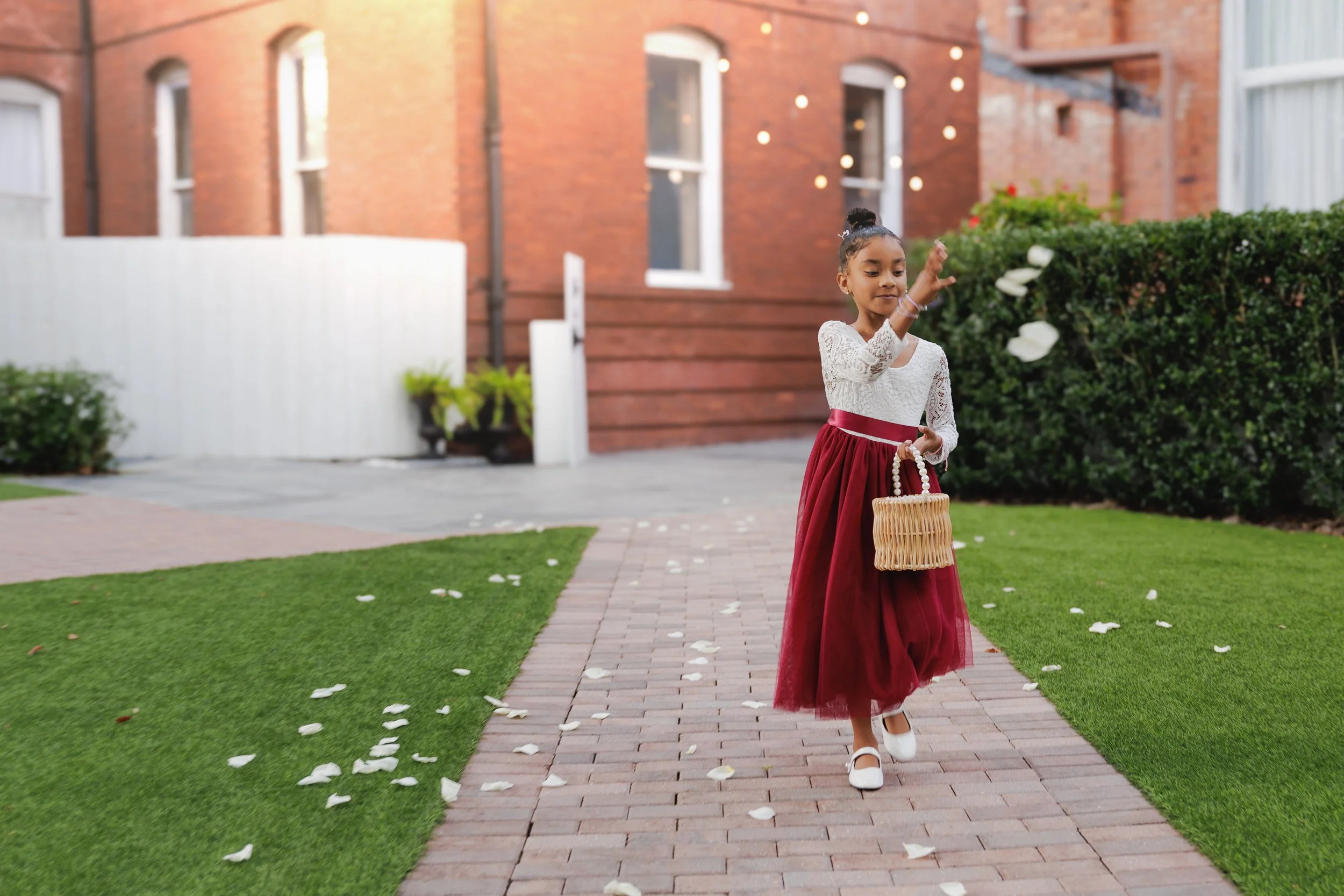 A young girl dressed in a white lace top and a long burgundy skirt walks on a brick sidewalk surrounded by green grass and flower petals, holding a wicker basket, as she throws flower petals.