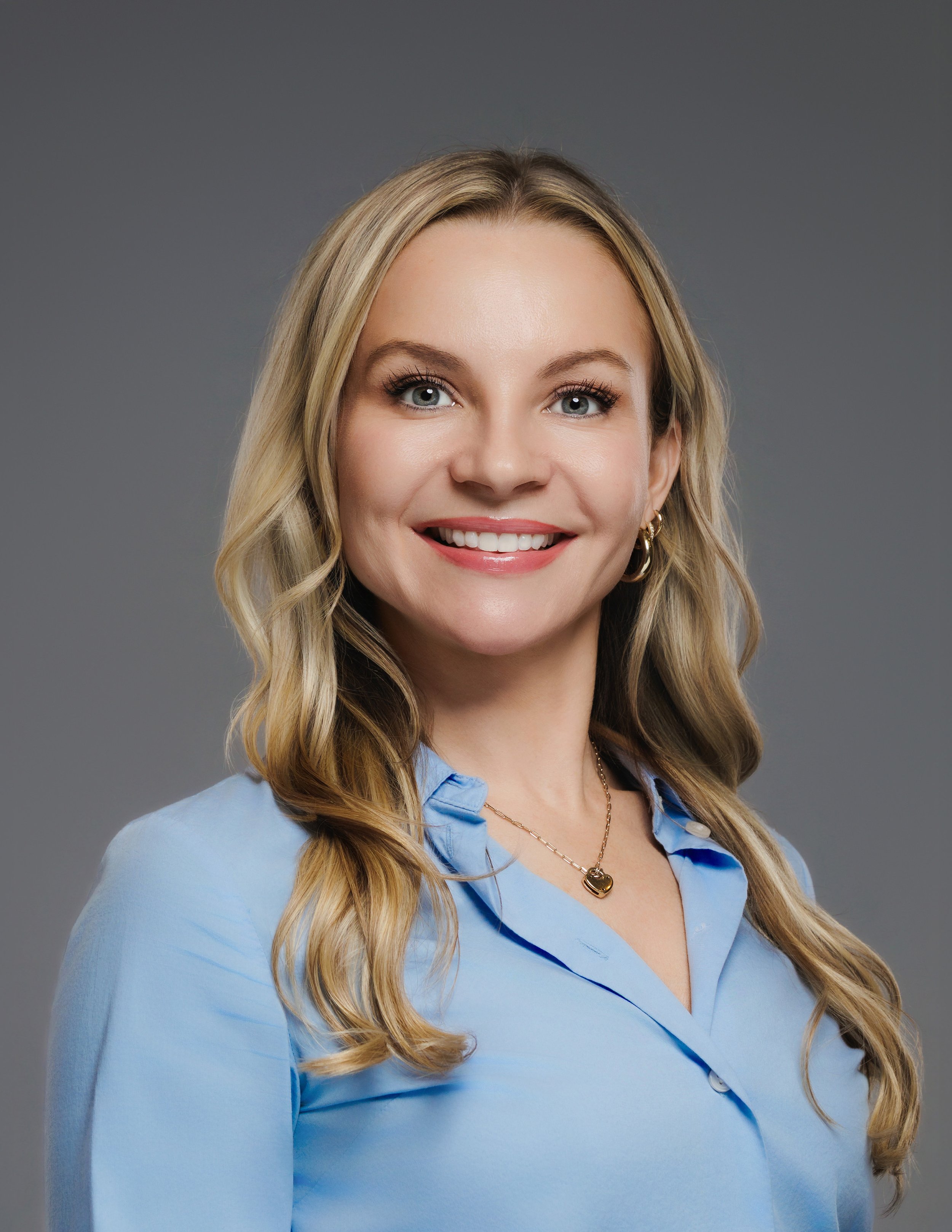 Portrait of a young woman with blonde hair wearing a light blue blouse and gold jewelry.