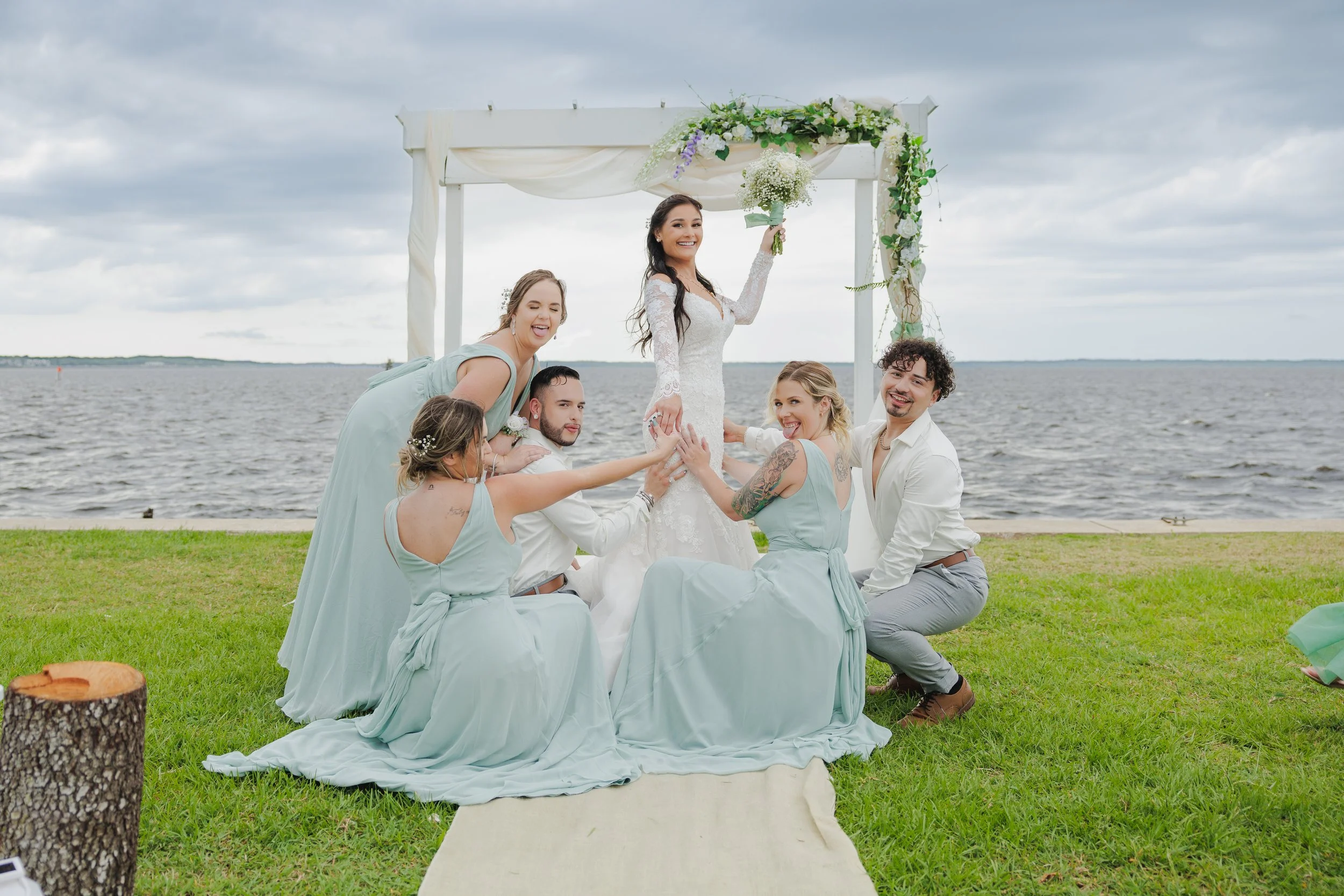 A wedding ceremony by a lake with a bride in a white lace gown holding a bouquet, surrounded by seven bridesmaids and groomsmen, all smiling and posing on a grassy area under a decorated arch.