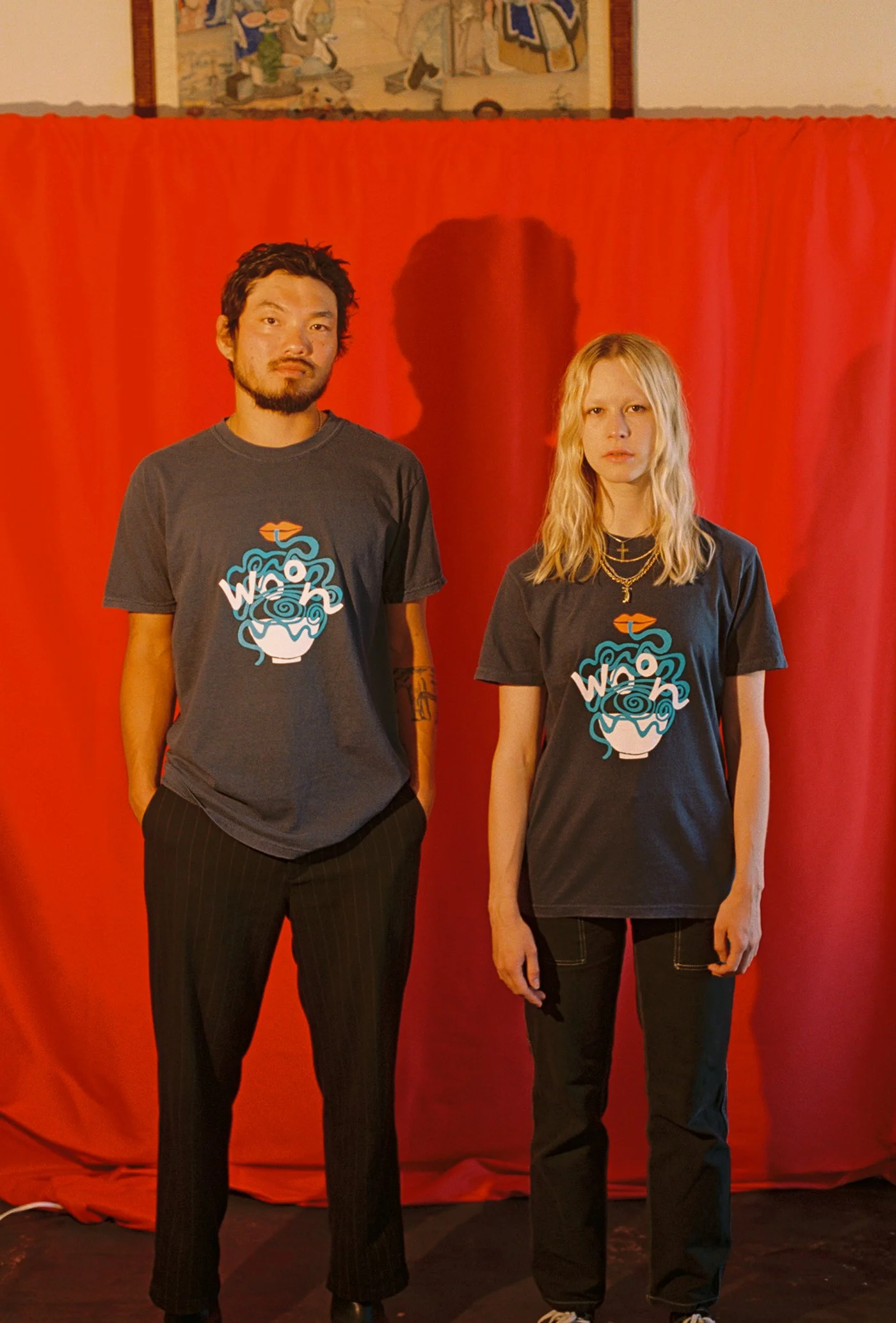 Two people wearing black t-shirts with red, white and blue artwork of a pair of lips eating noodles from a bowl