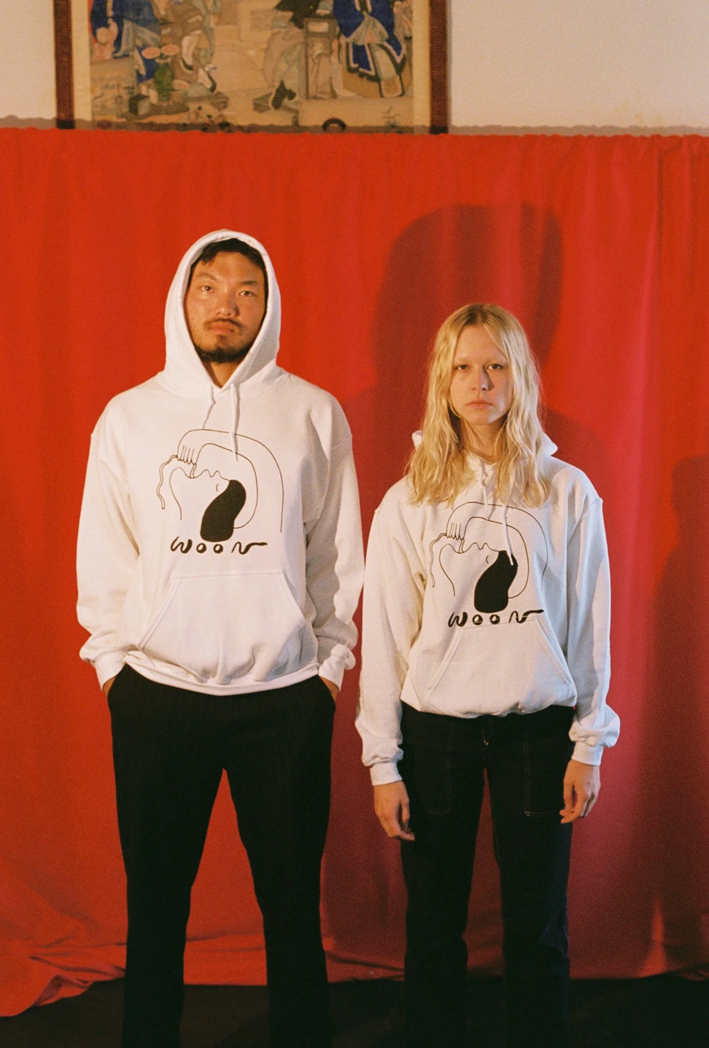 Two people wearing white hoodies with black artwork of a person eating a noodle