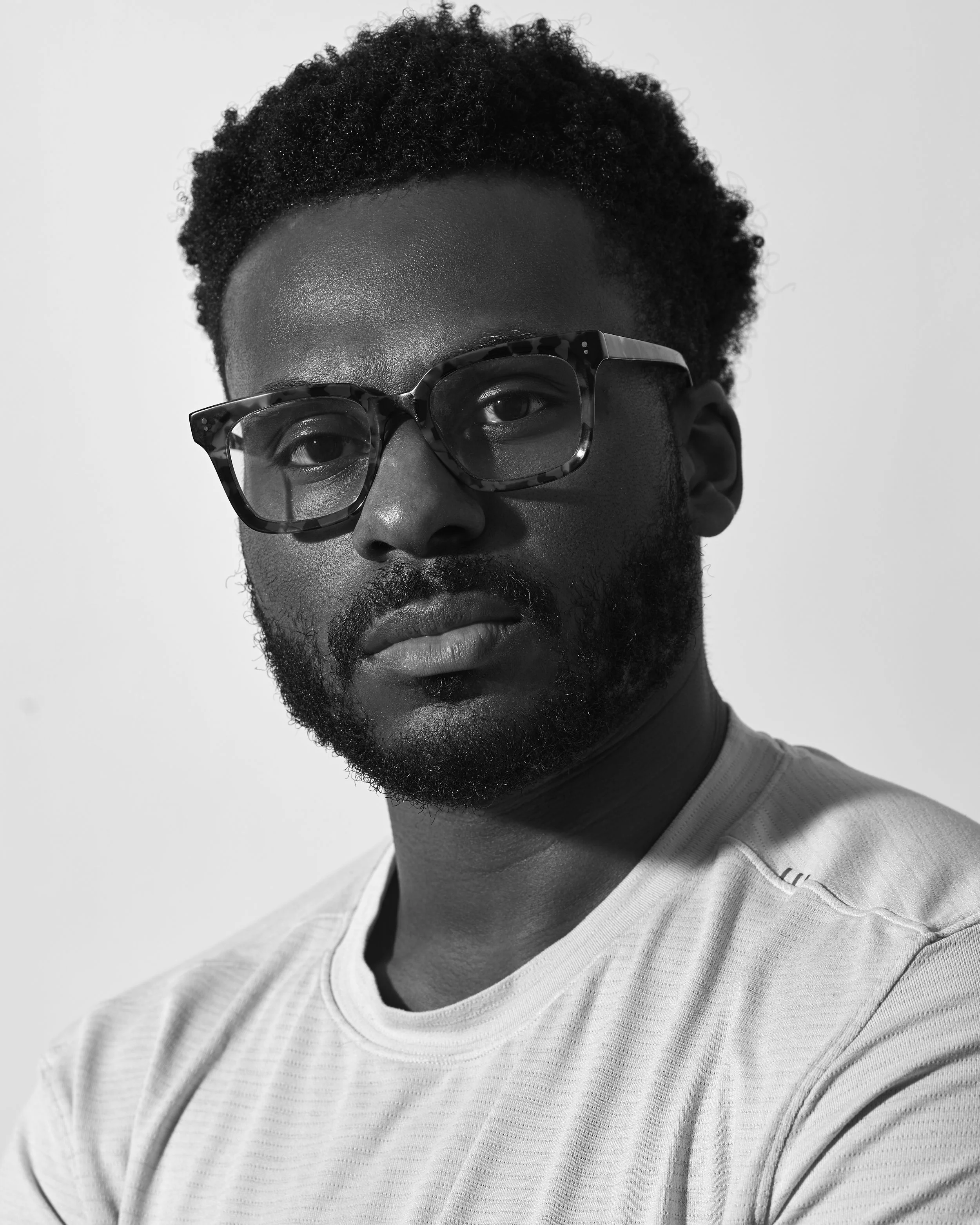 Black man with natural hair wearing black-framed glasses and a white textured shirt, looking at the camera, in black and white.