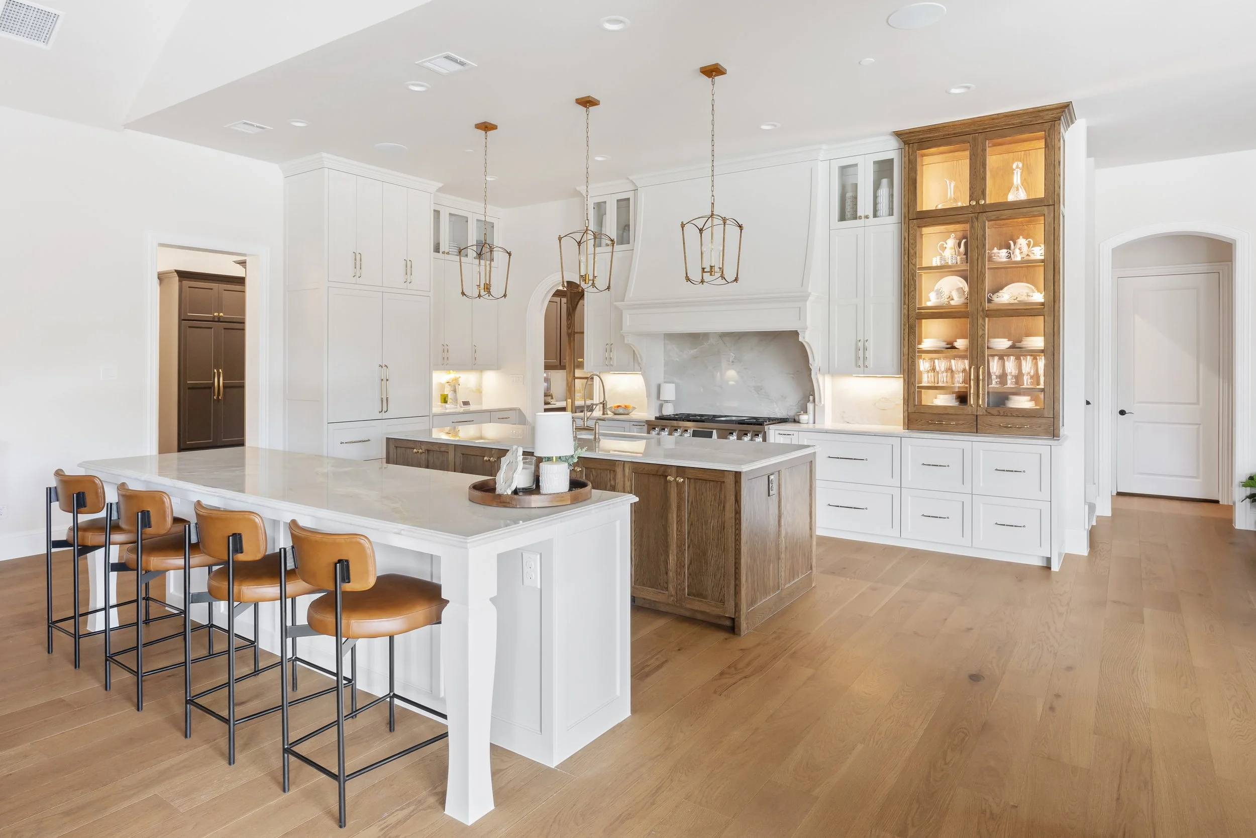 Spacious luxury kitchen with white cabinetry, wood island and wood hutch with quartzite countertops. Leather counter stools and brass pendants add warmth, while built in glass hutch cabinet displays fine dishwater, encasing the elegant design