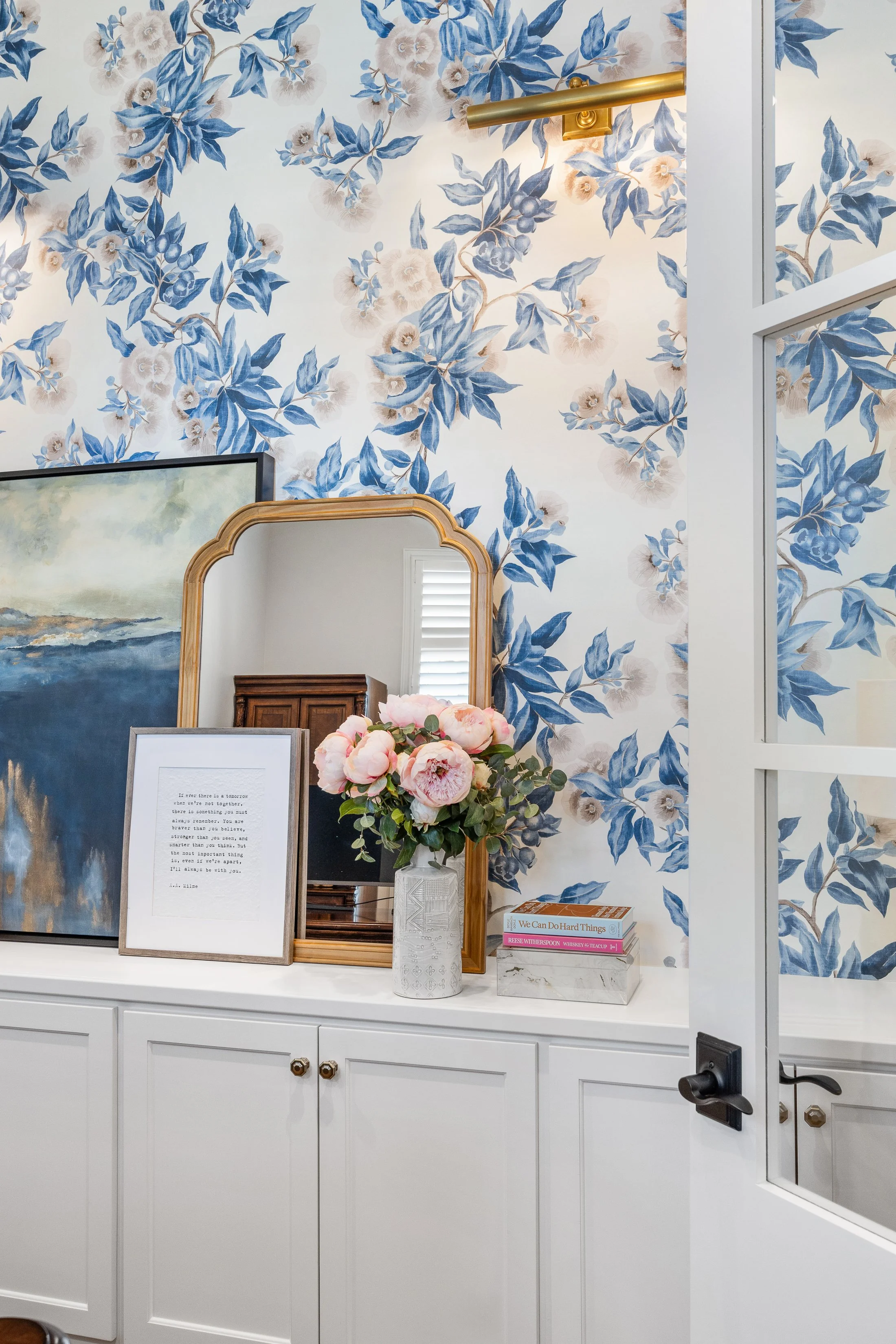 White built-in cabinetry styled with gold mirror, modern artwork, pink peonies and books. Blue floral wallpaper adds a bold, elegant backdrop to this transitional home office. Blending classic charm with contemporary details.