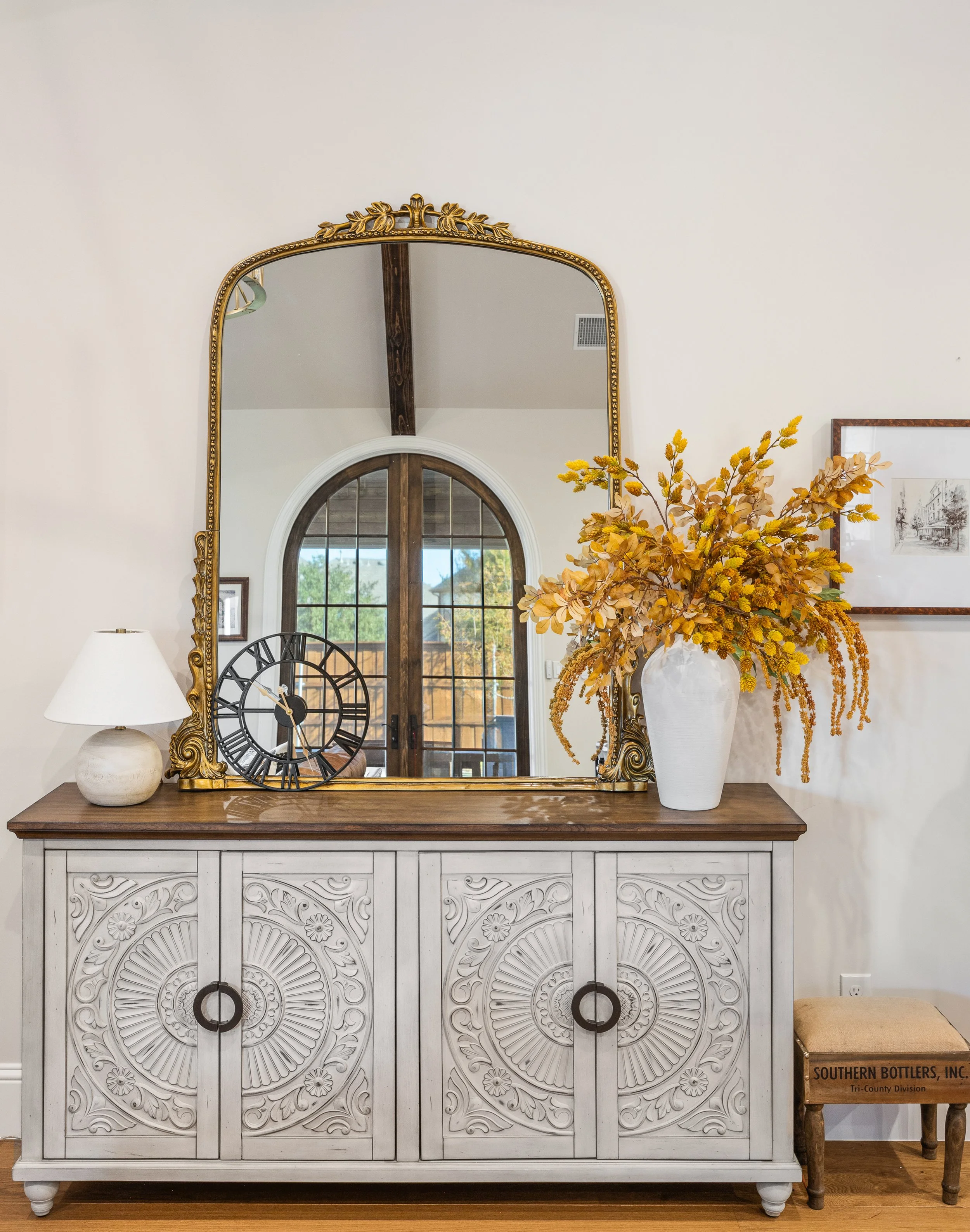 Carved wood sideboard with intricate carved detail, styled with large ornate gold mirror, ceramic table lamp, black clock and white vase with autumn foliage. Arched French door reflection shows off architecture of this custom home.