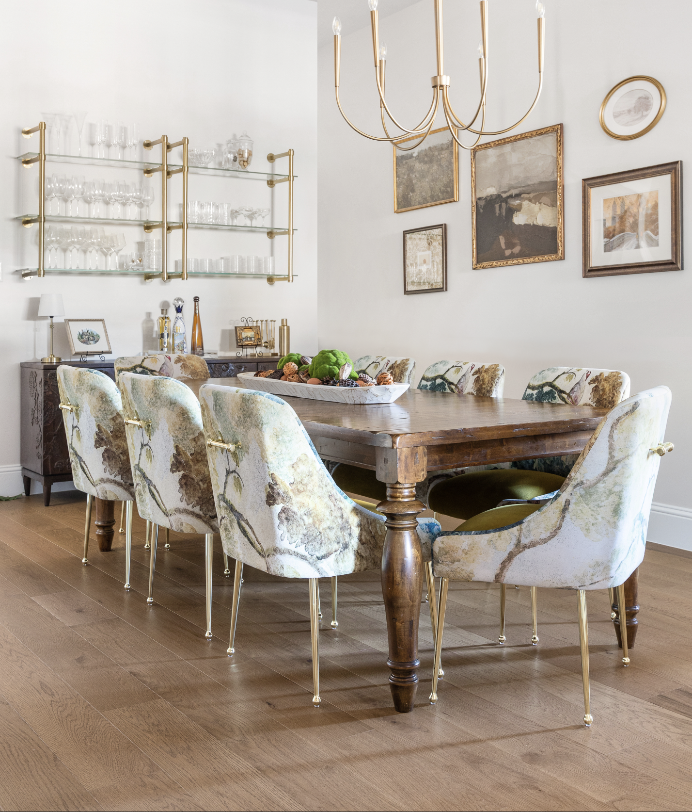 Refined dining room featuring a classic wood table paired with velvet floral-upholstered chairs and brass legs. A traditional gold chandelier hangs above, while glass shelving display crystal glassware. Traditional, neutral wall art and oak floors.