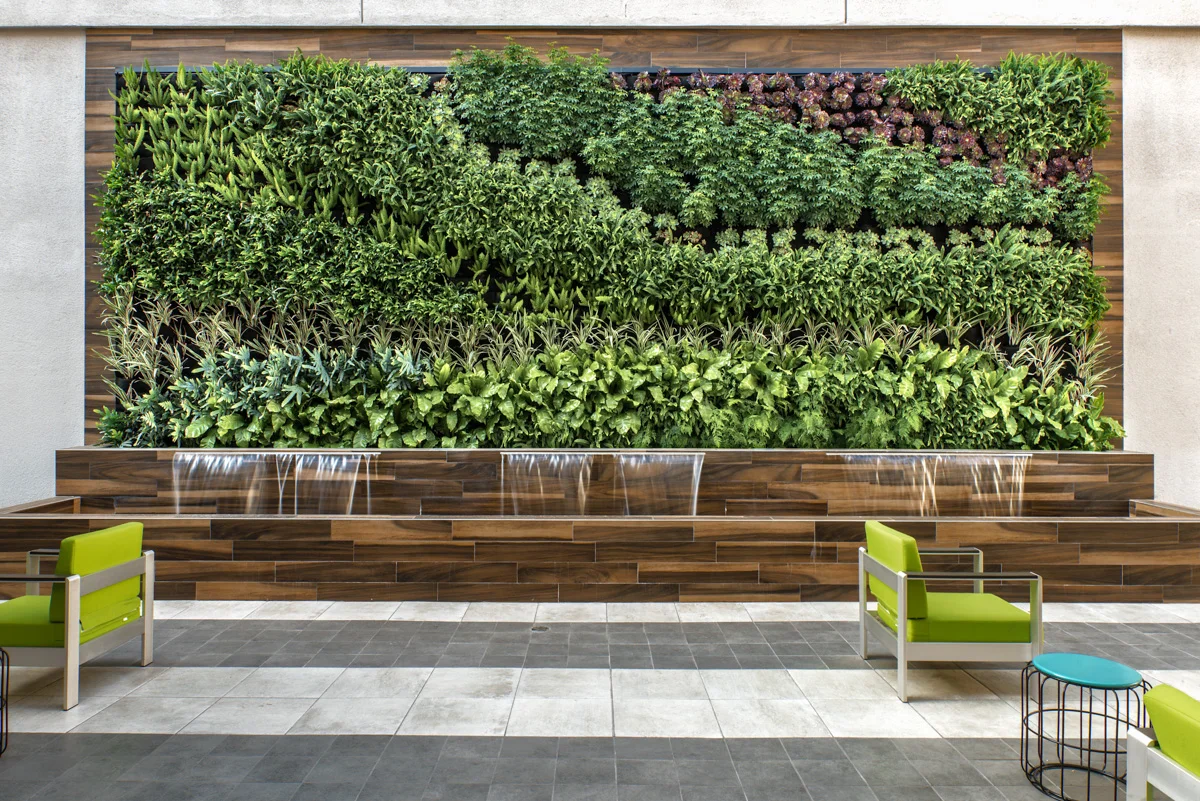 Indoor green wall with plants and water feature, with seating area including chairs with green cushions and a small table.