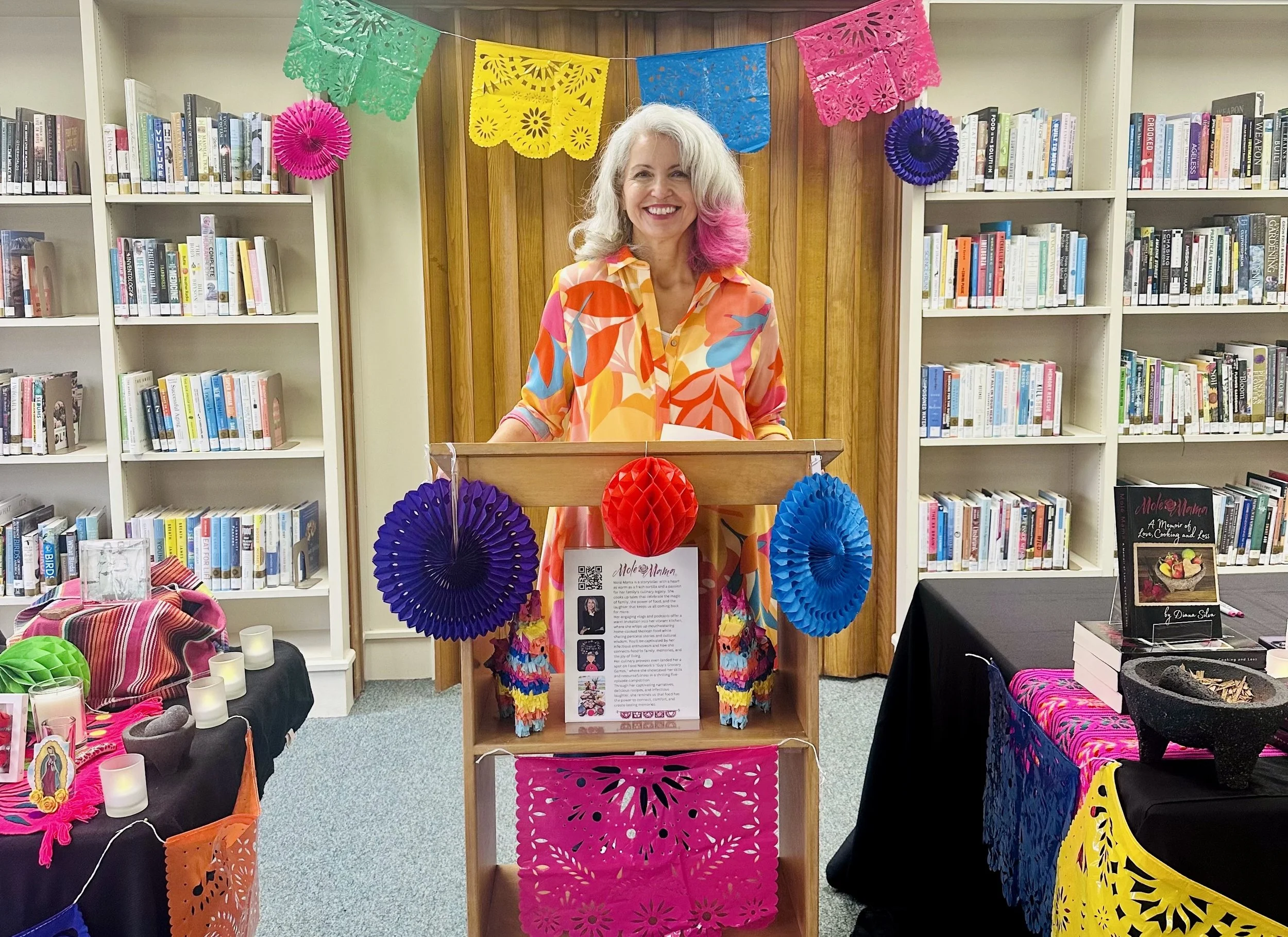 Molé Mama speaking at a library author event beside her books and traditional Mexican decorations.
