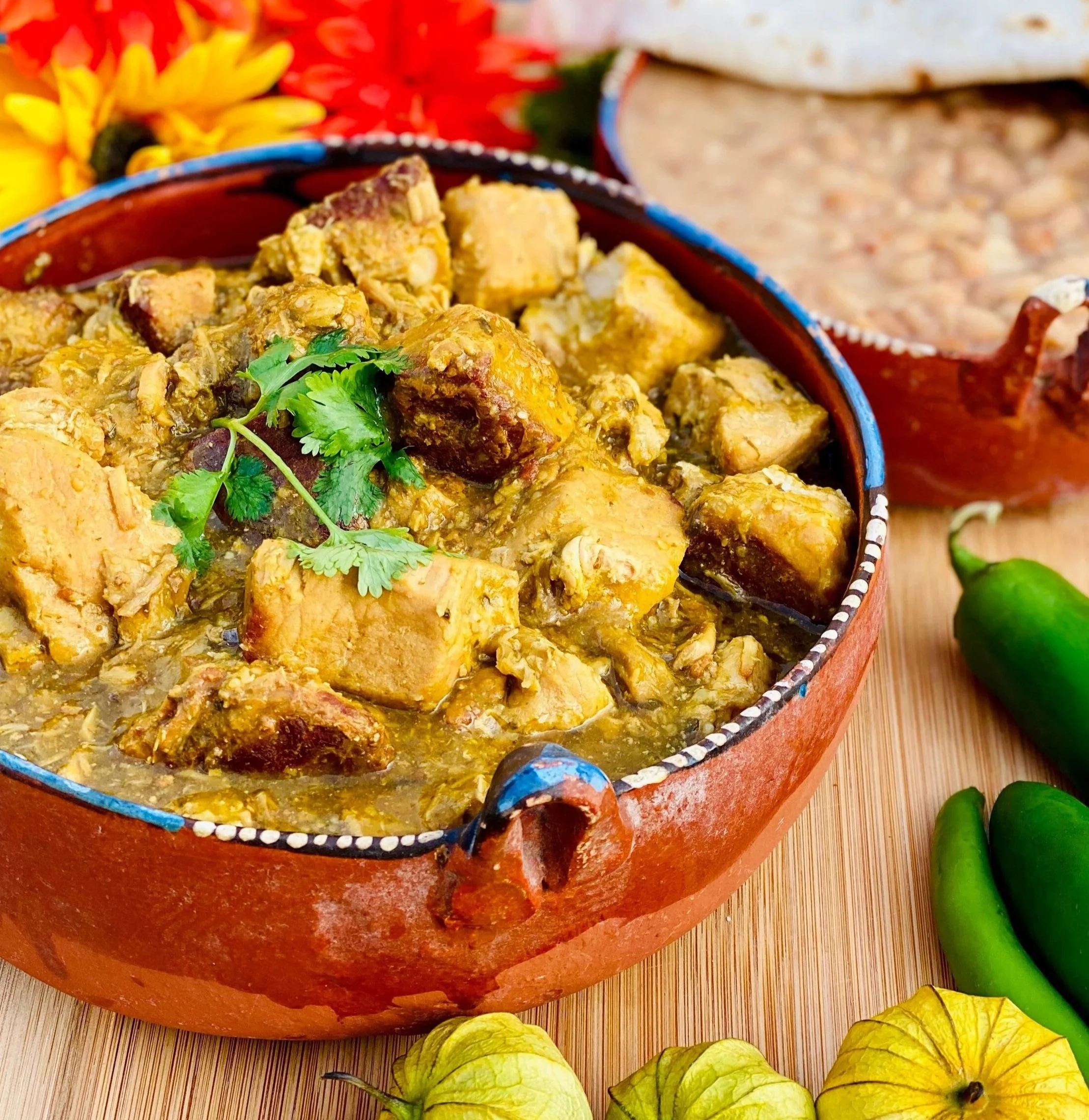 Chile Verde served in brown pottery bowl with refried beans, tortillas and bright flowers