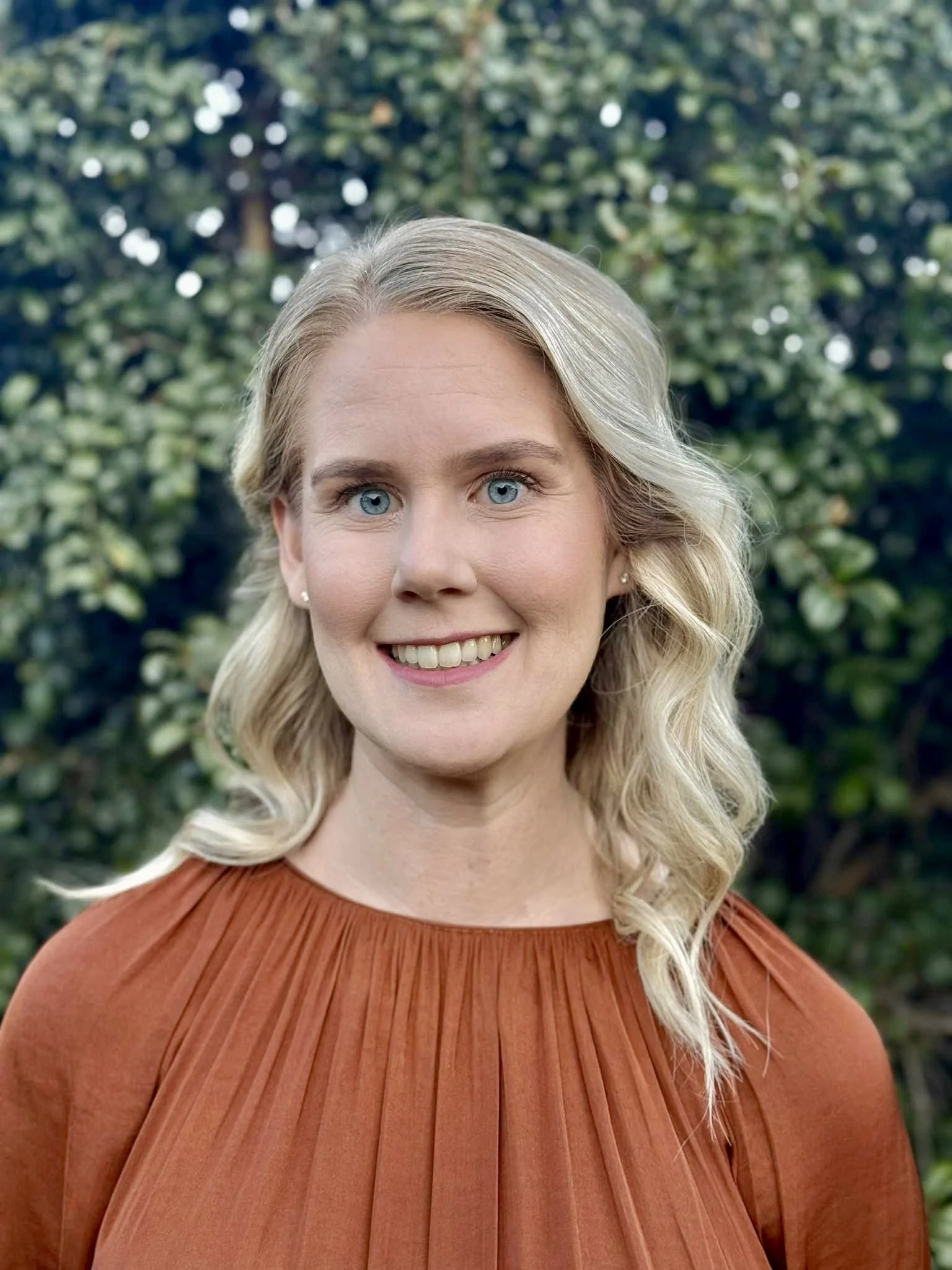 A woman with blonde, wavy hair and blue eyes smiling outdoors, wearing an orange pleated top with greenery in the background.