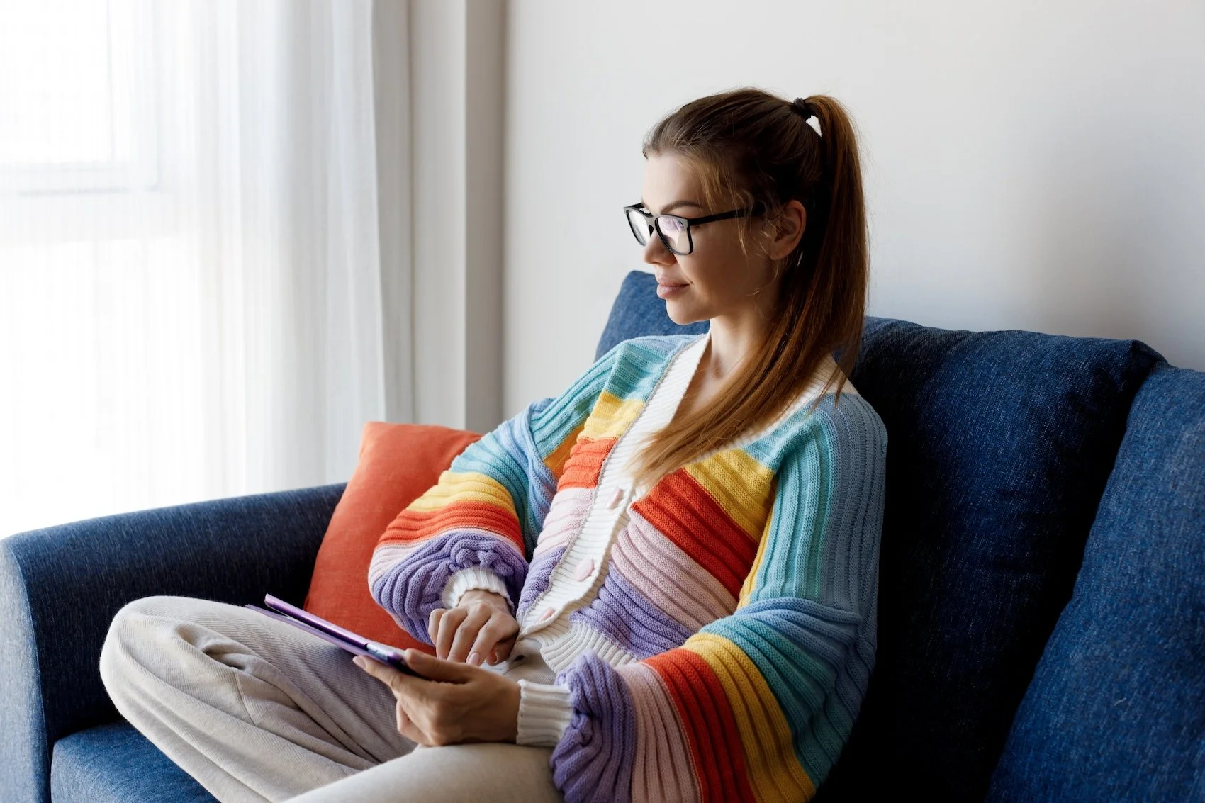 A woman with glasses and brown hair tied back in a ponytail, sitting on a dark blue couch, looking at a tablet device, wearing a colorful rainbow-striped sweater.