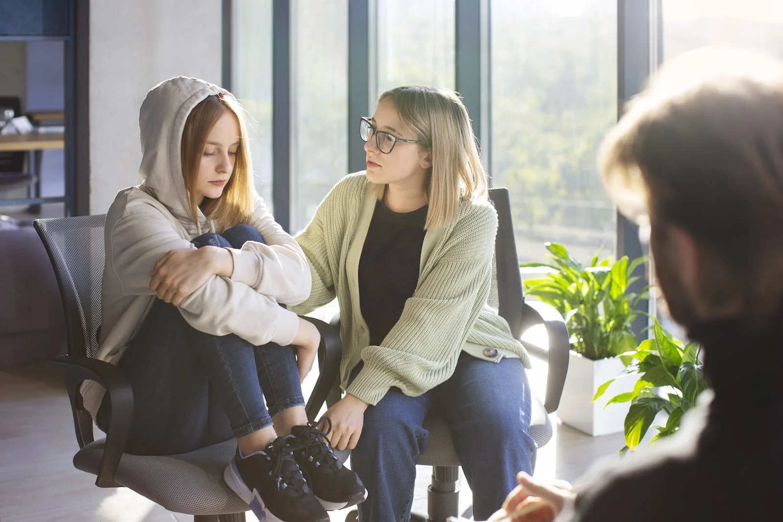 Woman comforting a girl with a hoodie during a therapy session in a bright room with large windows and plants.
