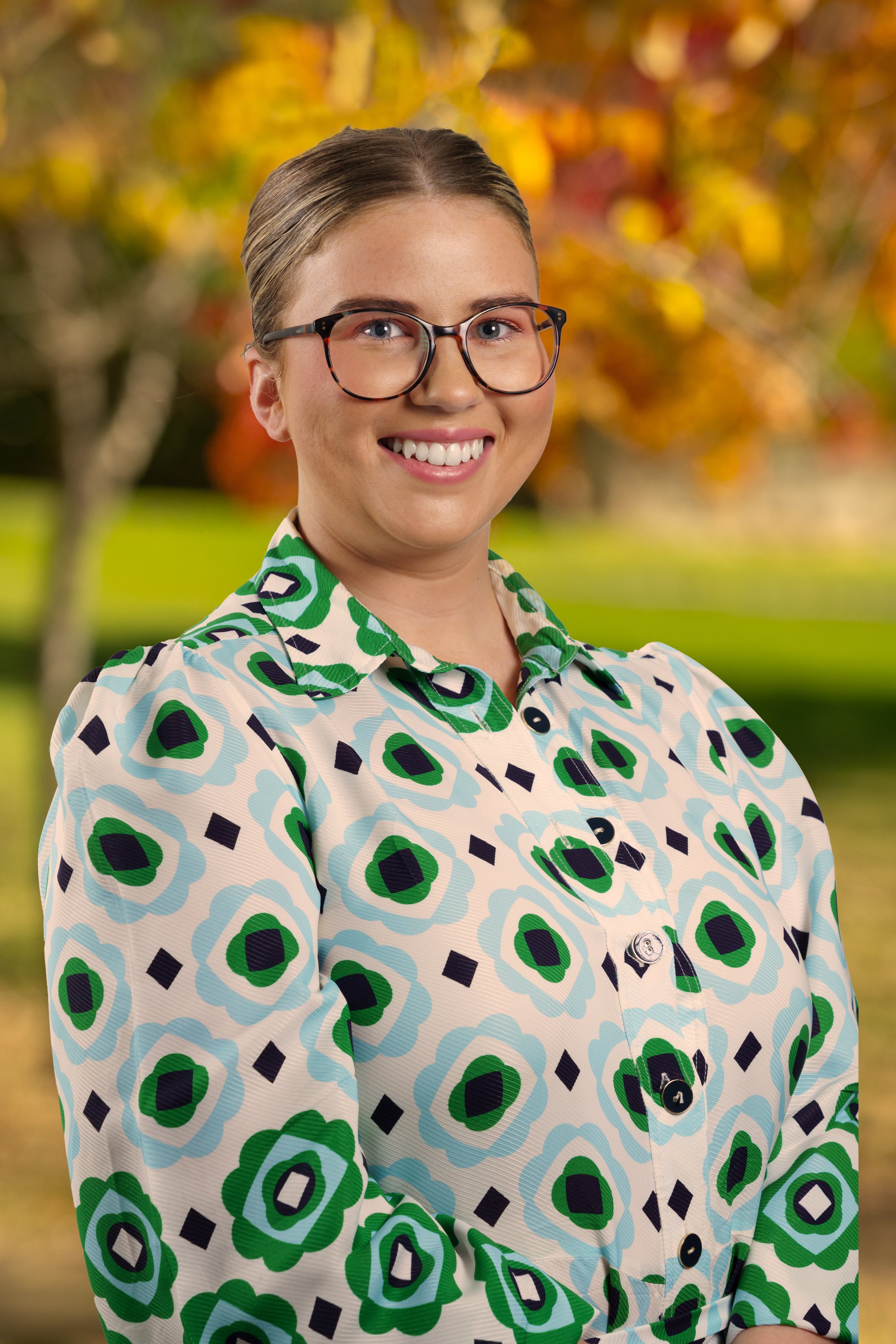 A smiling woman with glasses and her hair tied back, wearing a patterned blouse with green, blue, and black shapes, standing outdoors with autumn leaves in the background.