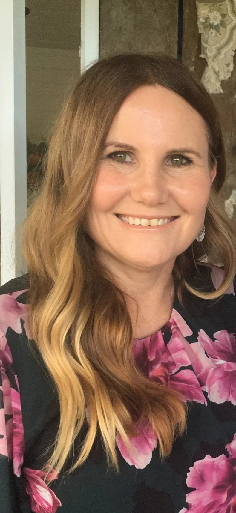 A woman with long, wavy, light brown hair, green eyes, smiling, wearing a black and pink floral dress, and silver earrings.
