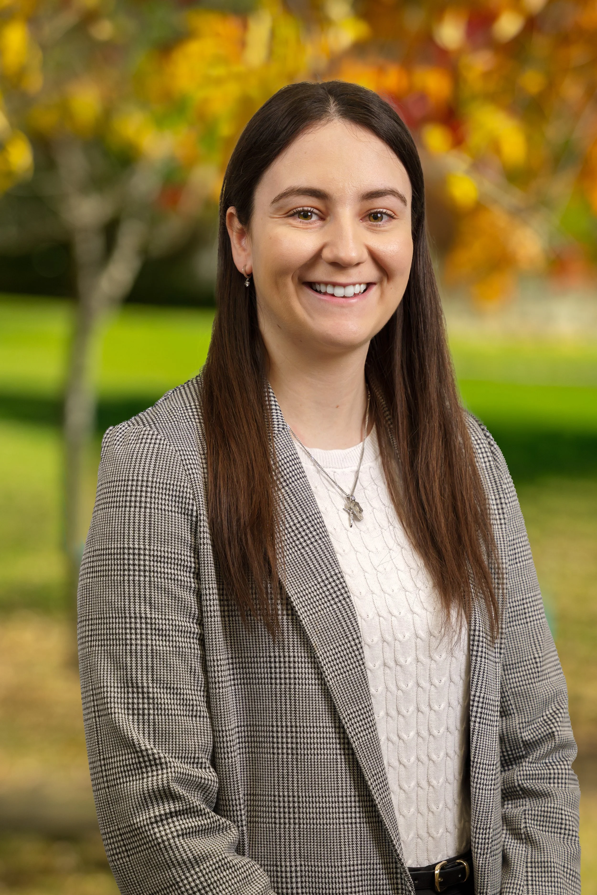 A smiling woman with long brown hair, wearing a plaid blazer and a white knit sweater, standing outdoors with autumn trees and fallen leaves in the background.