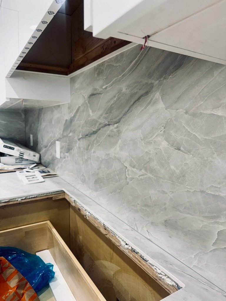 Close-up of a kitchen backsplash in progress, featuring a large gray marble tile with white and darker gray veins, with some construction materials and tools visible.