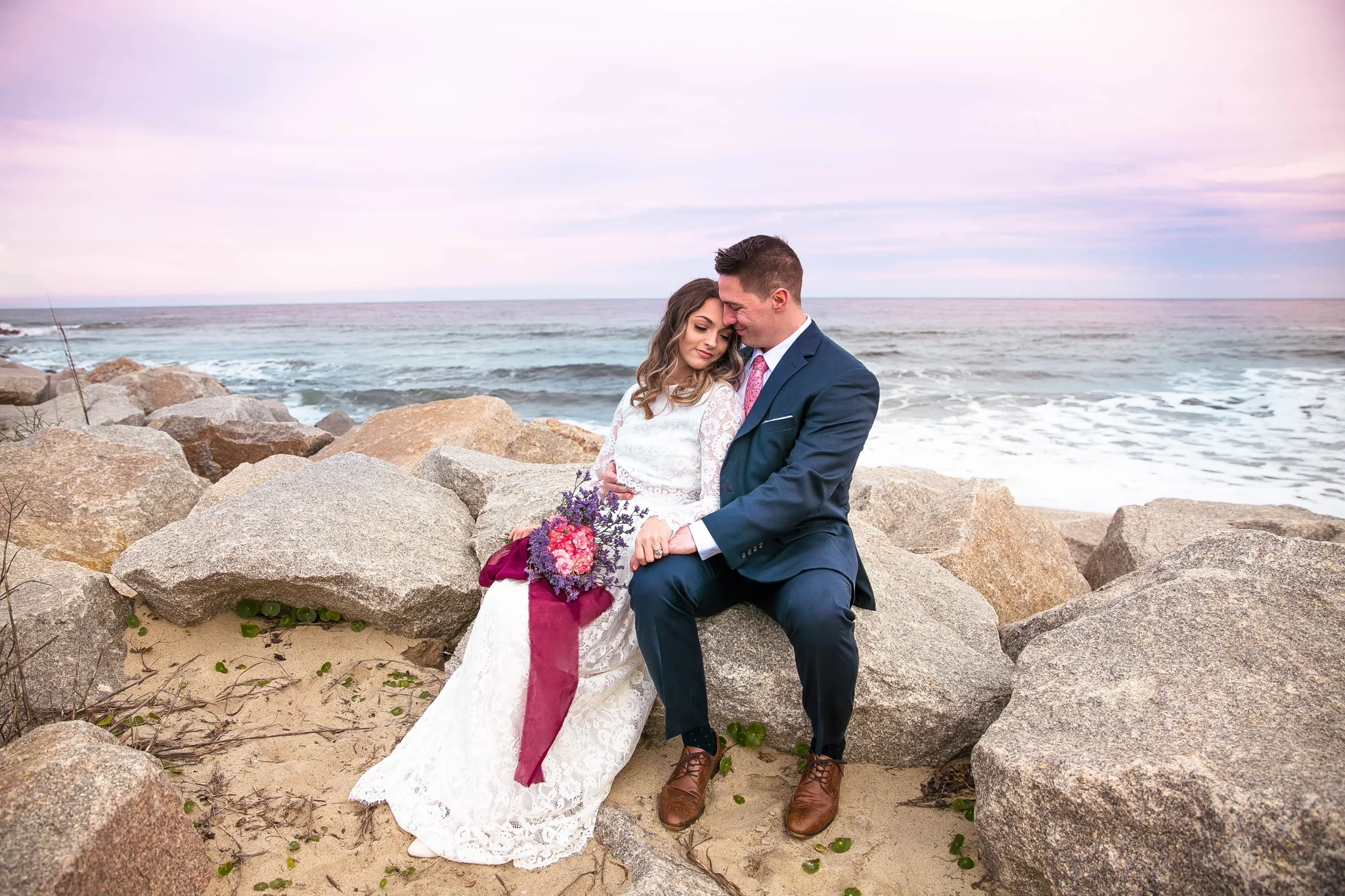 Cotton Candy Sky Beach Elopement Portraits - Bride and Groom sitting on top of rocks Cliffs - - dress by asos with purple and pink flowers and navy suit - oahu hawaii wedding photographer