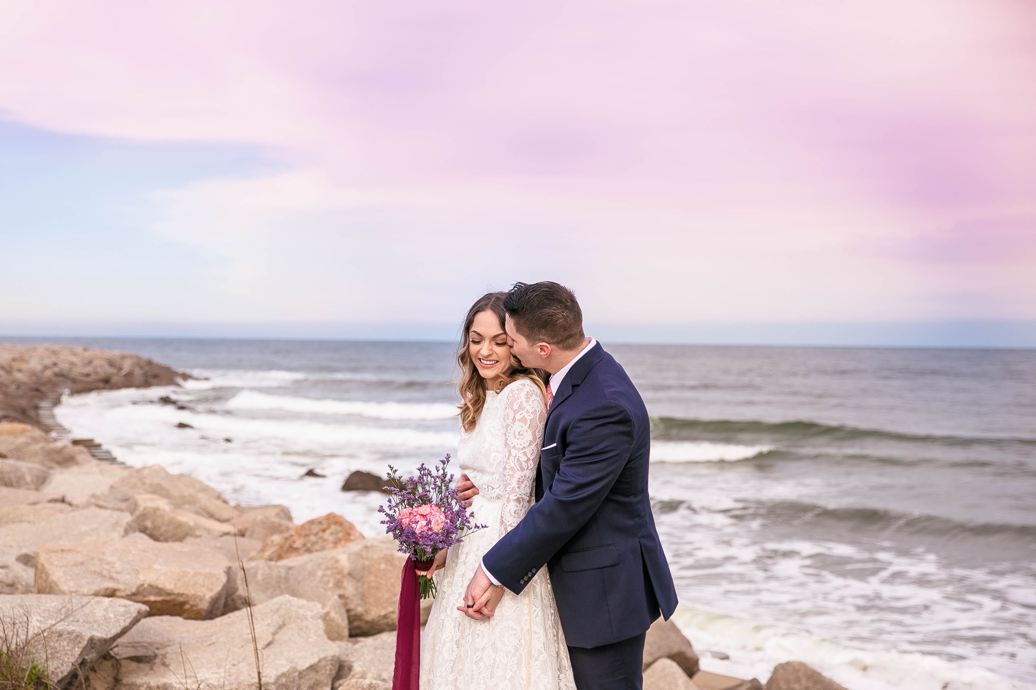Cotton Candy Sky Beach Elopement Portraits - Bride and Groom sitting on top of rocks Cliffs - - dress by asos with purple and pink flowers and navy suit - oahu hawaii wedding photographer