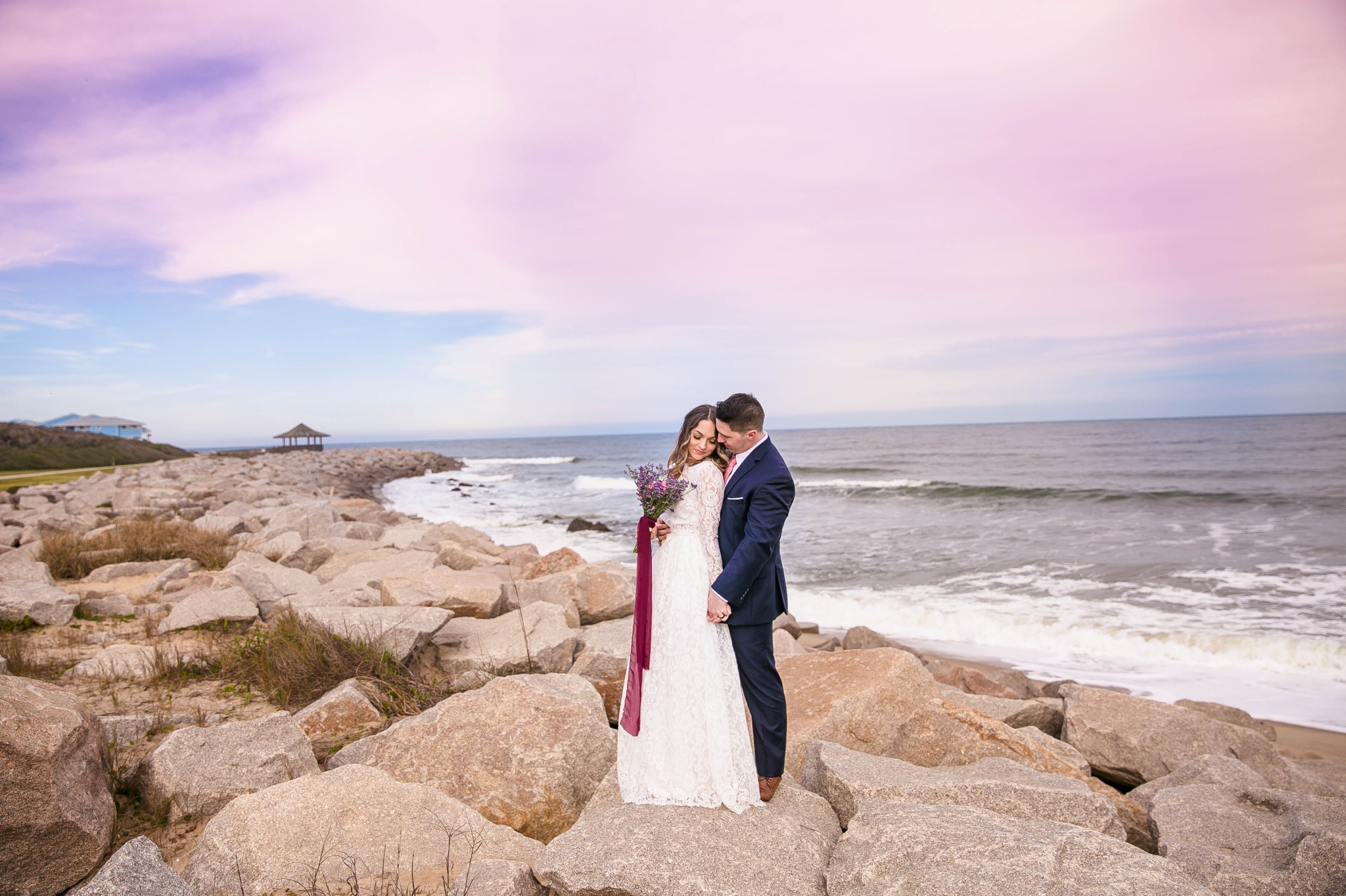 Cotton Candy Sky Beach Elopement Portraits - Bride and Groom sitting on top of rocks Cliffs - - dress by asos with purple and pink flowers and navy suit - oahu hawaii wedding photographer