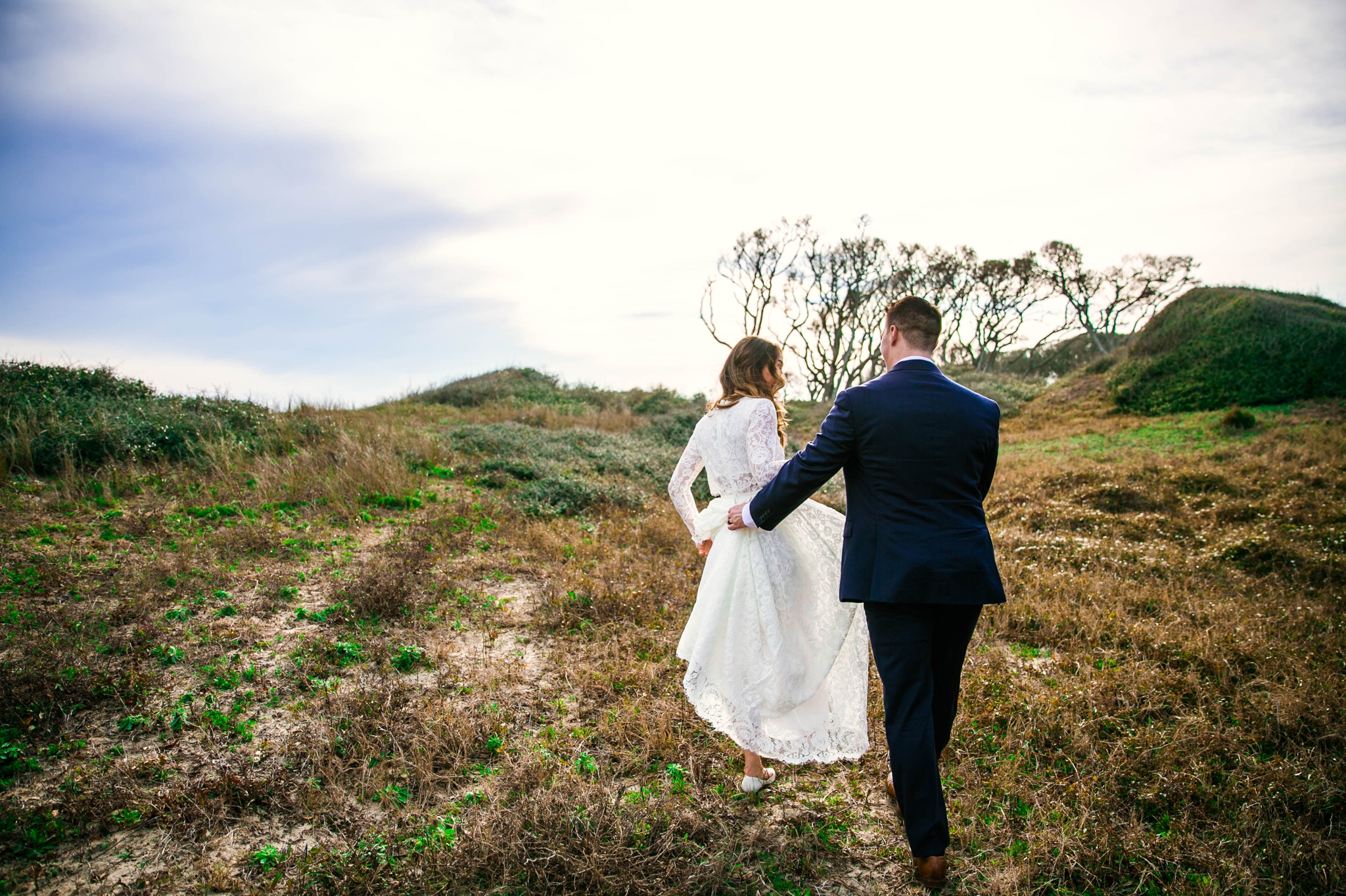 Groom holding brides dress while walking in Lush Green Hills - Beach Elopement Photography - wedding dress by asos with purple and pink flowers and navy suit - oahu hawaii wedding photographer