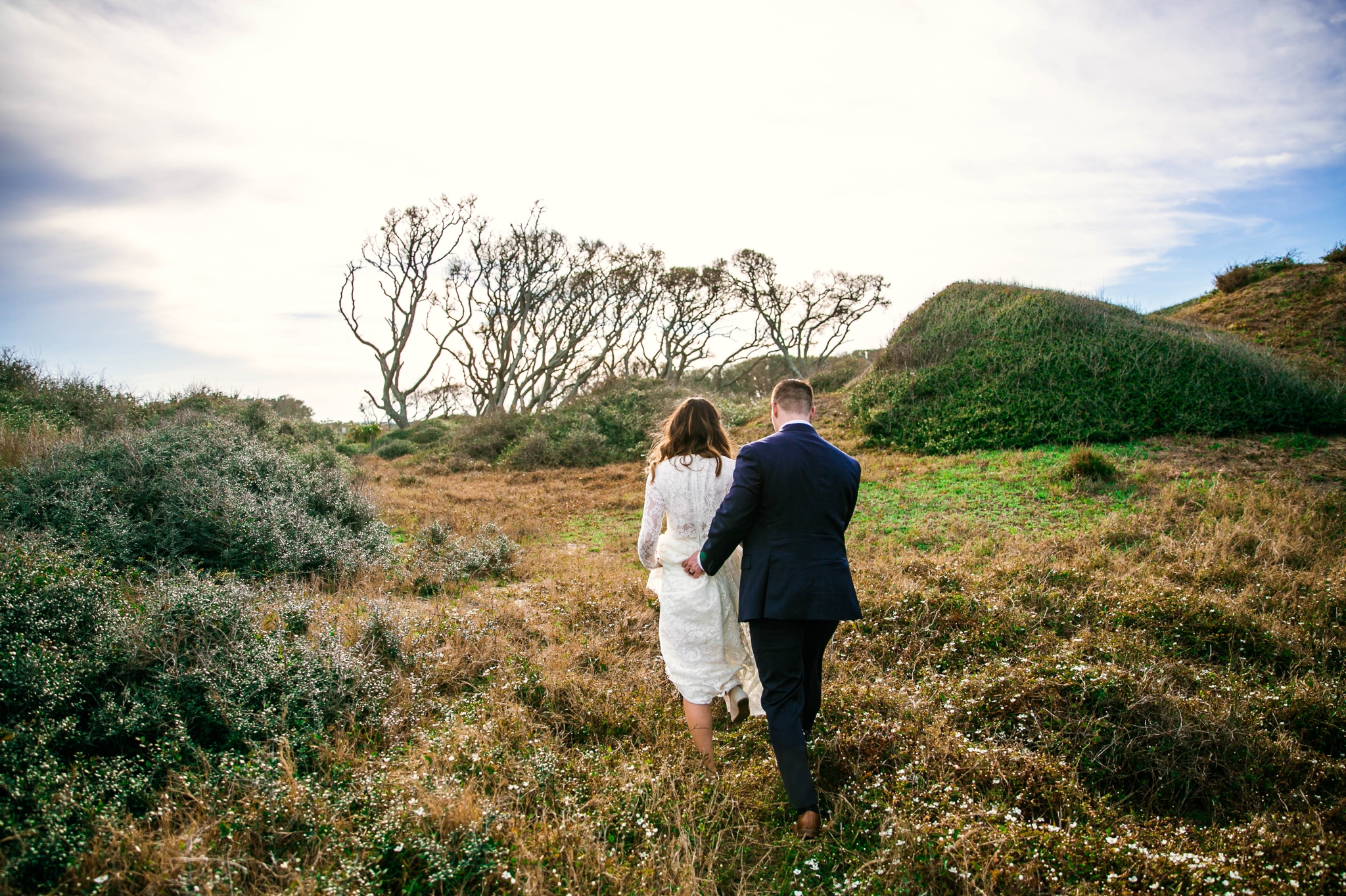 Groom holding brides dress while walking in Lush Green Hills - Beach Elopement Photography - wedding dress by asos with purple and pink flowers and navy suit - oahu hawaii wedding photographer