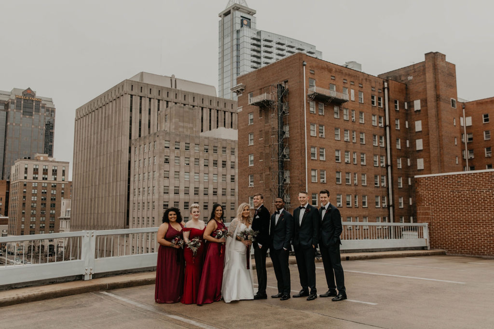 Downtown Wedding Party Portraits on top of a parking garage with the skyline in the background - Bride in an Allure Bridals Mermaid Gown - Burgundy Bridesmaids dresses by davids bridal Matt Smarsh and Johanna Dye - suites by the black tux - Raleigh North Carolina Urban Edgy Downtown Wedding - suits by the black tux