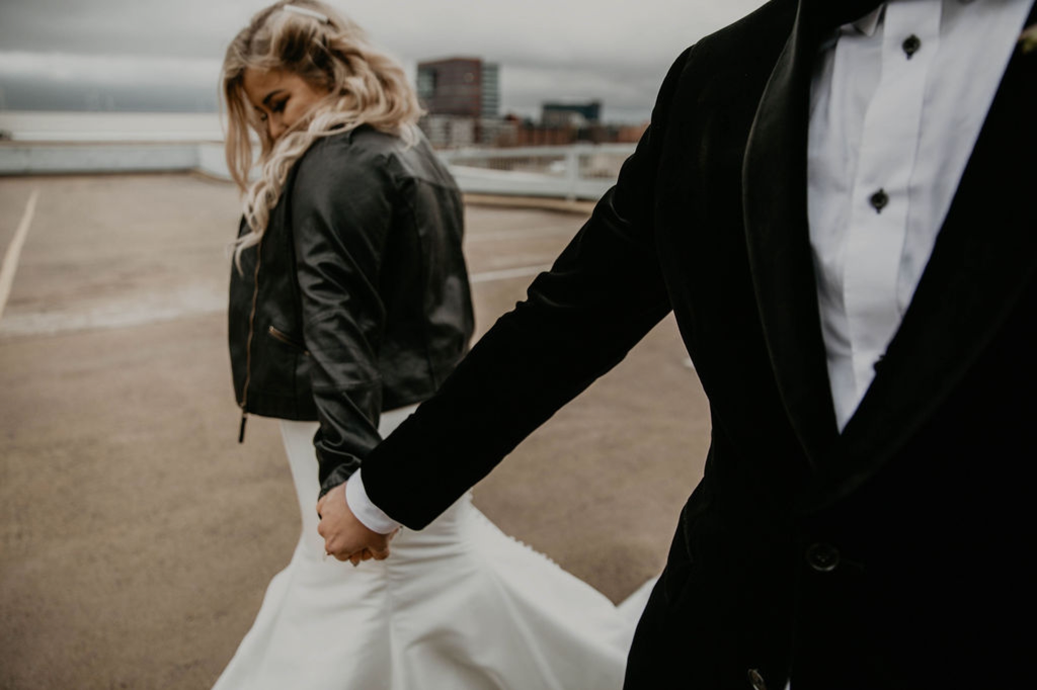 bride and groom hand in hand - Wedding Portraits in front of the raleigh skyline - - Bride in an Allure Bridals Mermaid Gown and a leather jacket - Burgundy Bridesmaids dresses by davids bridal Matt Smarsh and Johanna Dye - suites by the black tux - Raleigh North Carolina Urban Downtown Wedding - suits by the black tux