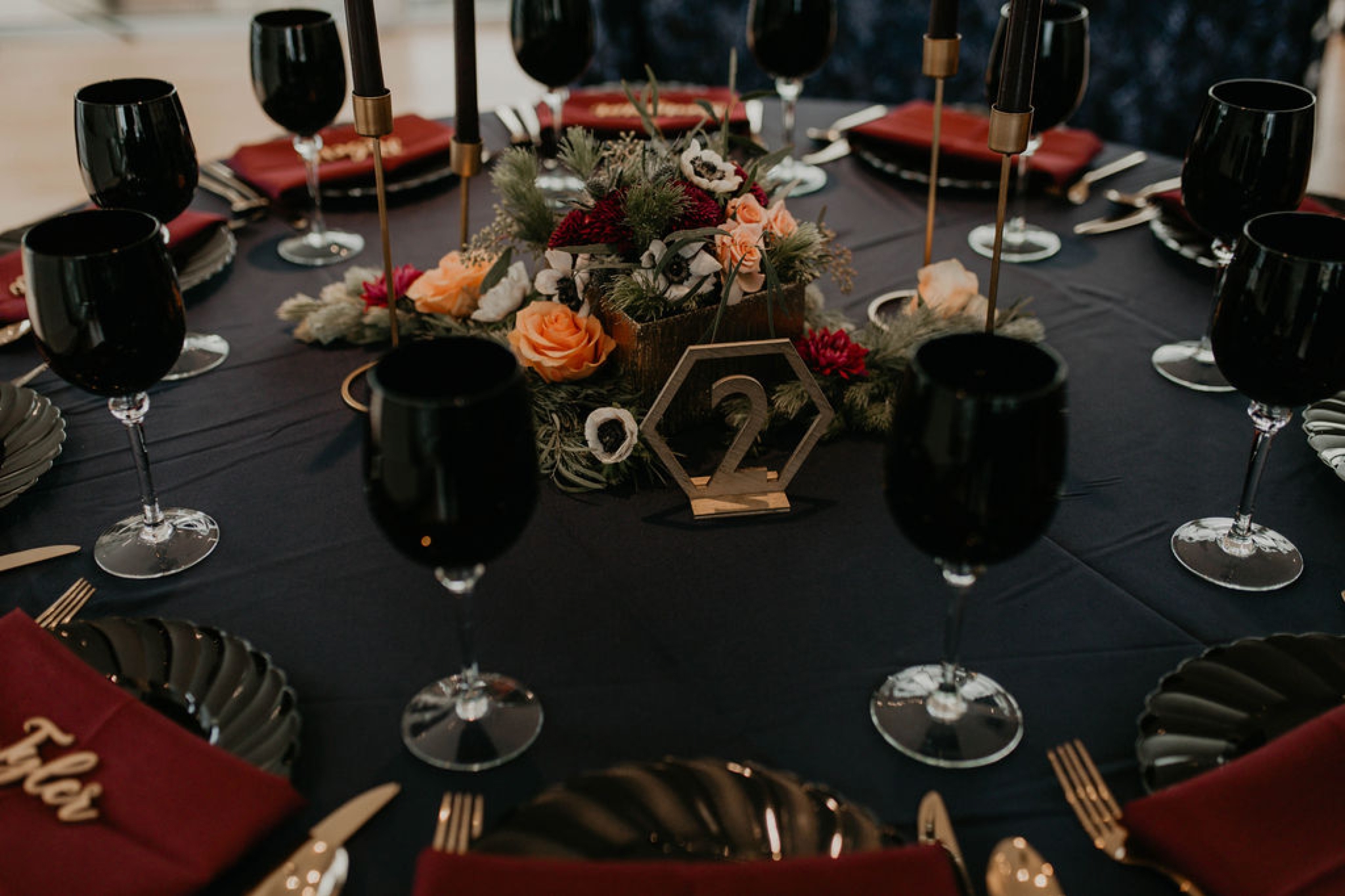 dark wedding table decorations - Navy tablecloth with burgundy napkins and gold laser cut name place cards, gold silverwear and black plates and black wine glasses by ce rental - dark flowers centerpiece - gold table number - gold candle stick holder with black candles - Matt Smarsh and Johanna Dye - Raleigh North Carolina Urban Downtown Wedding at the Glass Box at 230