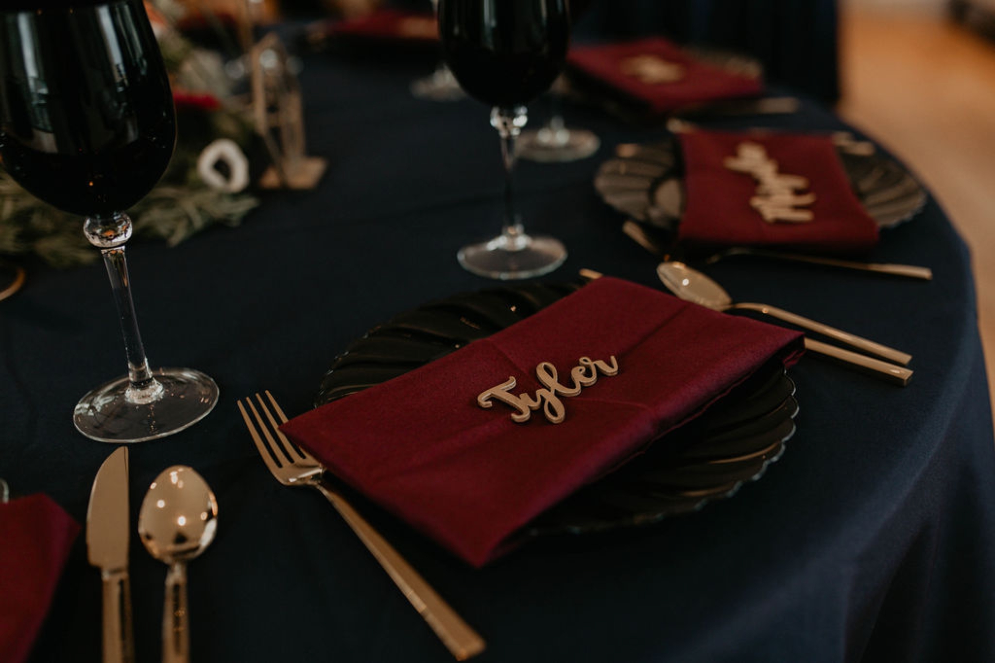 sweetheart table - dark wedding table decorations - Navy tablecloth with burgundy napkins and gold laser cut name place cards, gold silverwear and black plates and black wine glasses by ce rental - dark flowers centerpiece - gold table number - gold candle stick holder with black candles - Matt Smarsh and Johanna Dye - Raleigh North Carolina Urban Downtown Wedding at the Glass Box at 230