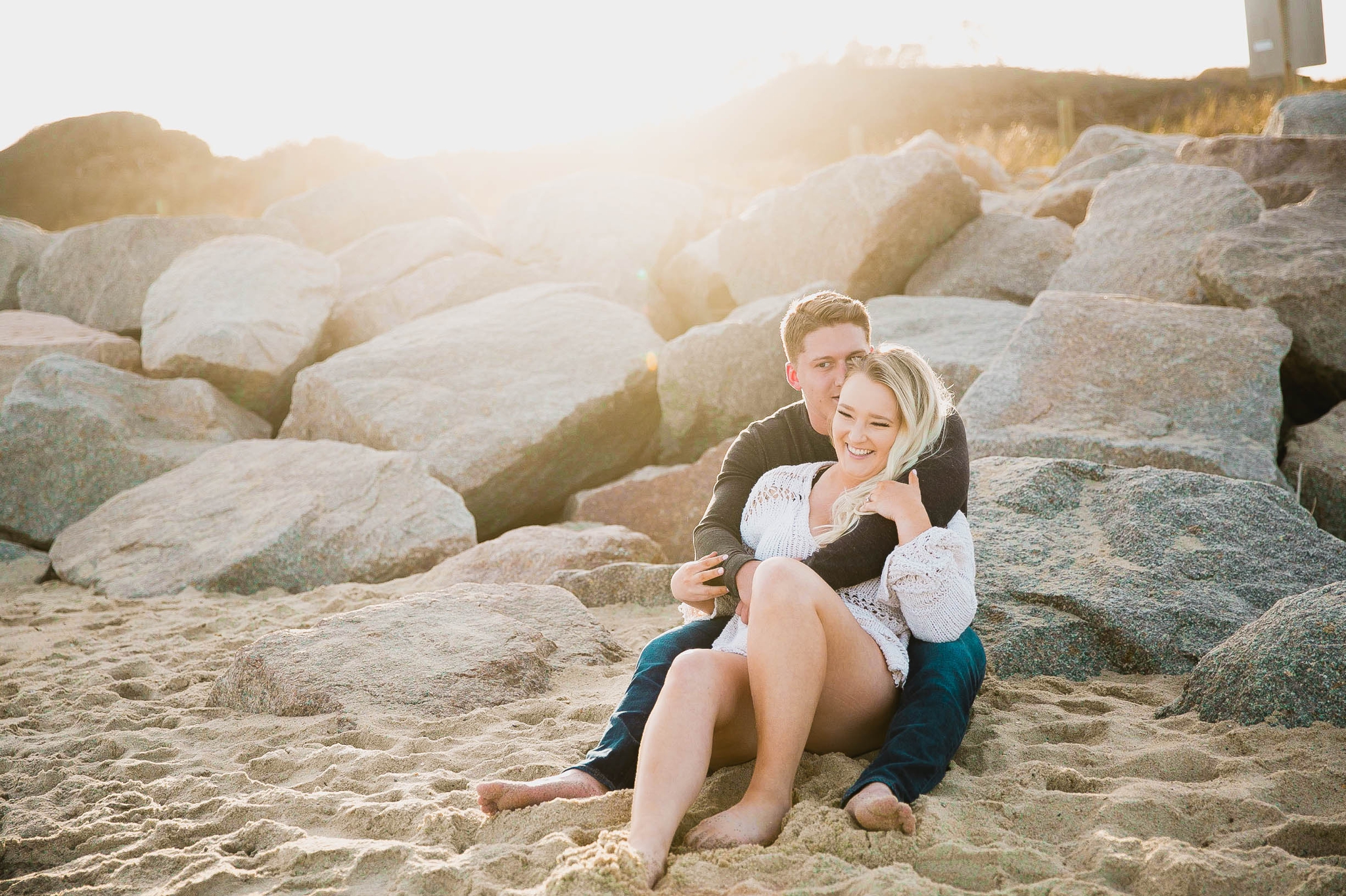  couple looking at the ocean while sitting on a sandy beach at sunset with golden hour light shining onto them - girl wearing ripped jeans shorts and a white free people sweater - Casual Beach Engagement Photography Session - Honolulu Oahu Hawaii Wed