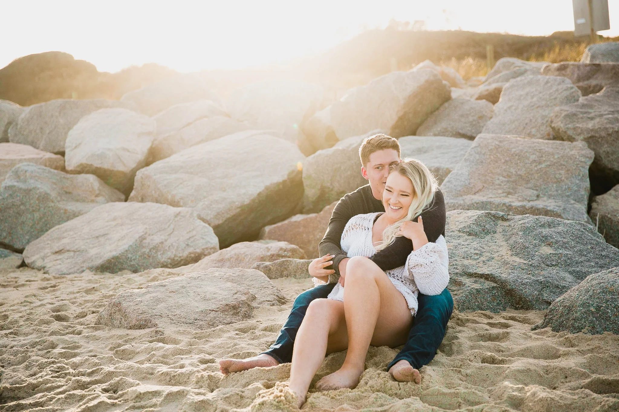 couple looking at the ocean while sitting on a sandy beach at sunset with golden hour light shining onto them - girl wearing ripped jeans shorts and a white free people sweater - Casual Beach Engagement Photography Session - Honolulu Oahu Hawaii Wedding Photographer - Johanna Dye