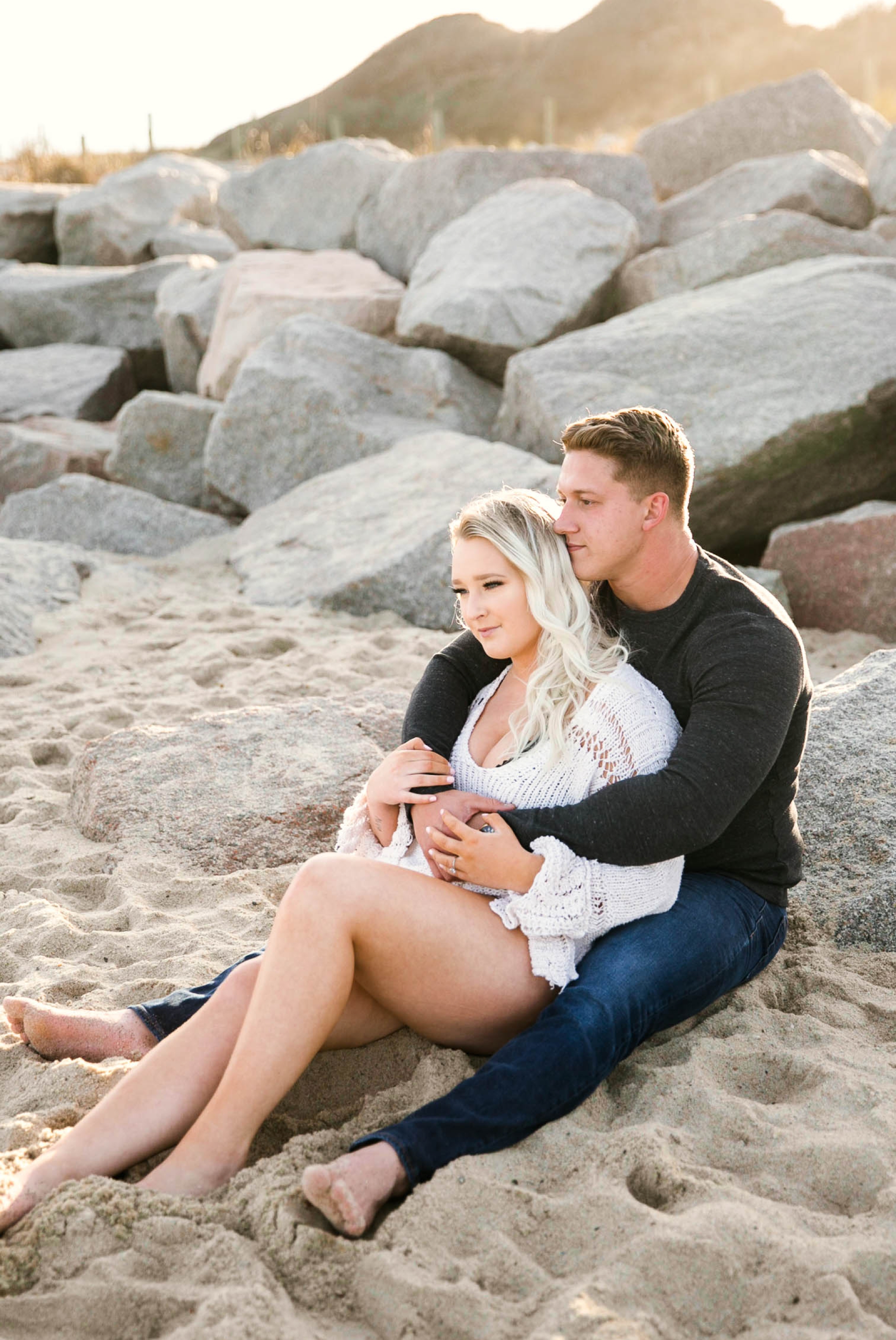  couple looking at the ocean while sitting on a sandy beach at sunset with golden hour light shining onto them - girl wearing ripped jeans shorts and a white free people sweater - Casual Beach Engagement Photography Session - Honolulu Oahu Hawaii Wed