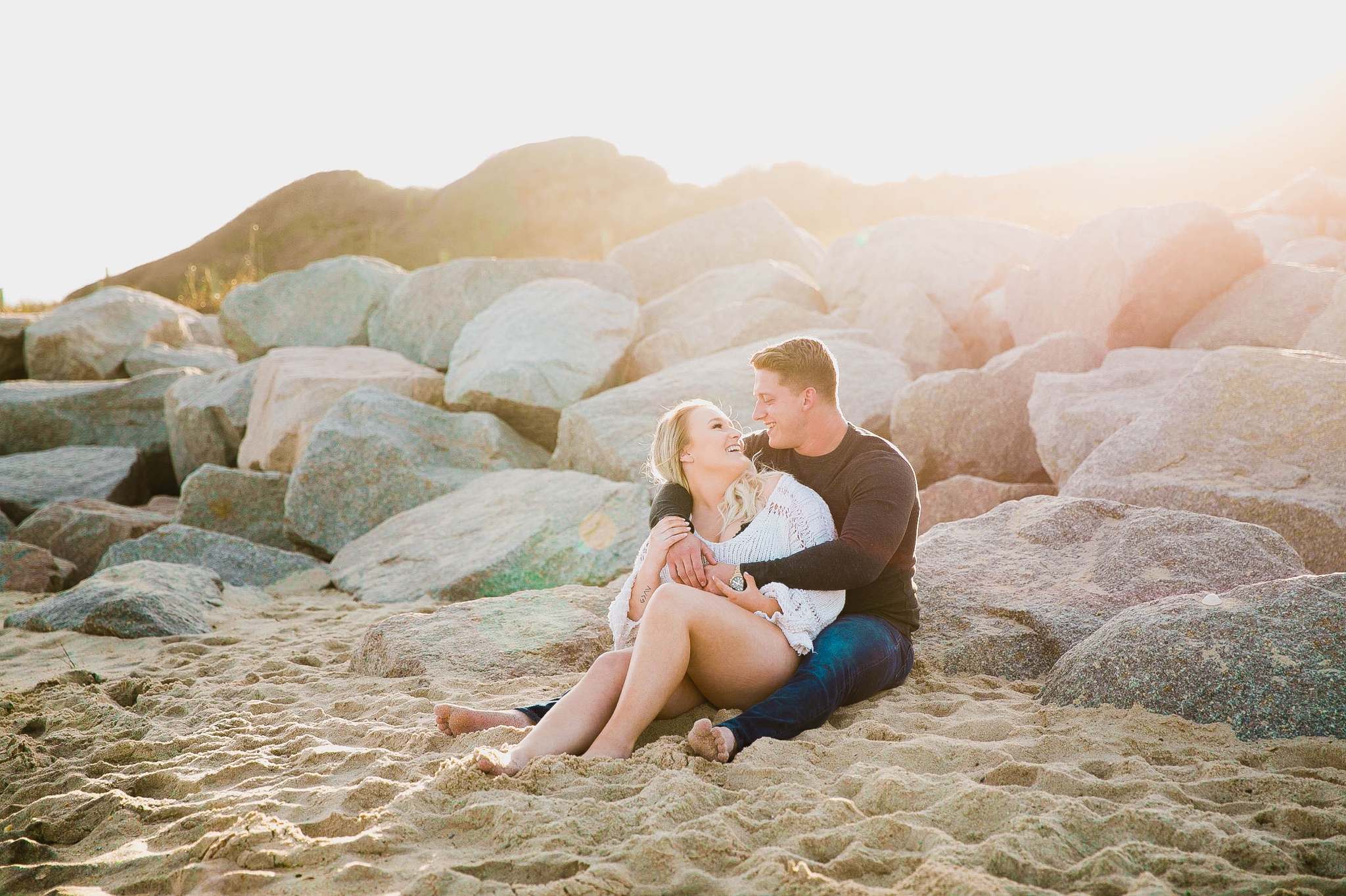  couple looking at the ocean while sitting on a sandy beach at sunset with golden hour light shining onto them - girl wearing ripped jeans shorts and a white free people sweater - Casual Beach Engagement Photography Session - Honolulu Oahu Hawaii Wed