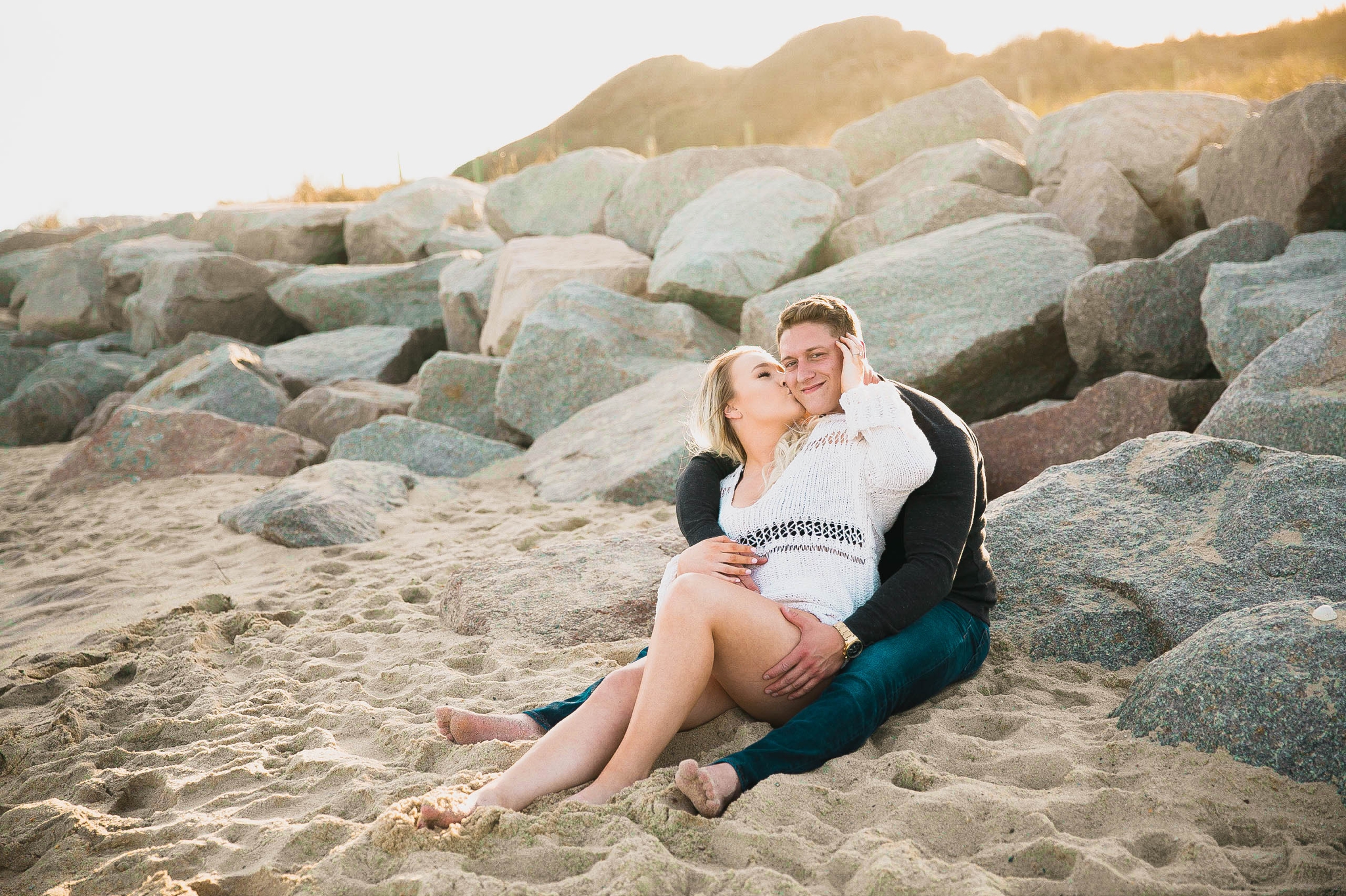  couple looking at the ocean while sitting on a sandy beach at sunset with golden hour light shining onto them - girl wearing ripped jeans shorts and a white free people sweater - Casual Beach Engagement Photography Session - Honolulu Oahu Hawaii Wed