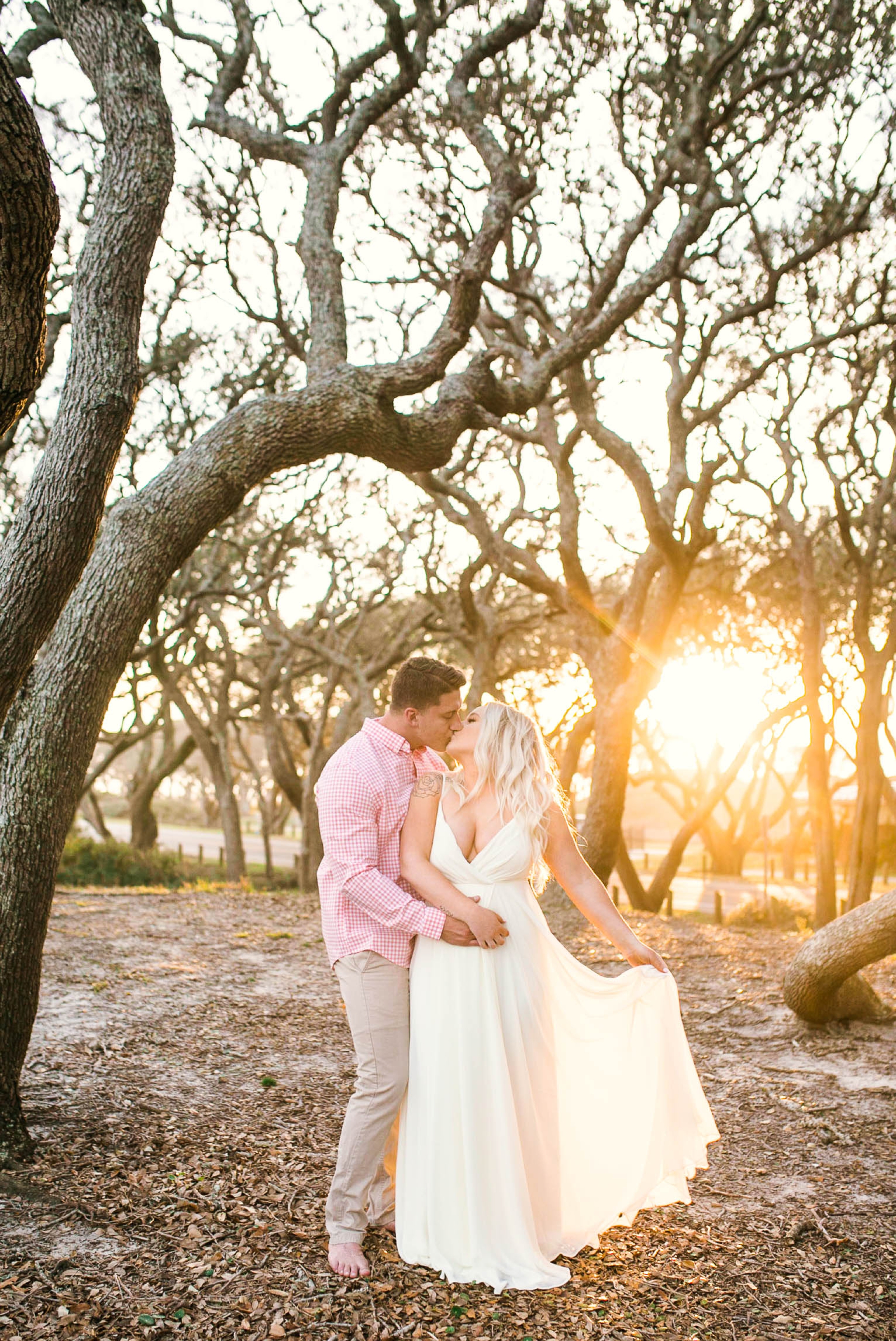  Engagement Photography Session beneath tropical trees at sunset during golden hour light - man kissing his fiance, woman is playing with her dress - girl is wearing a white flowy maxi dress from lulus - Honolulu Oahu Hawaii Wedding Photographer - Jo