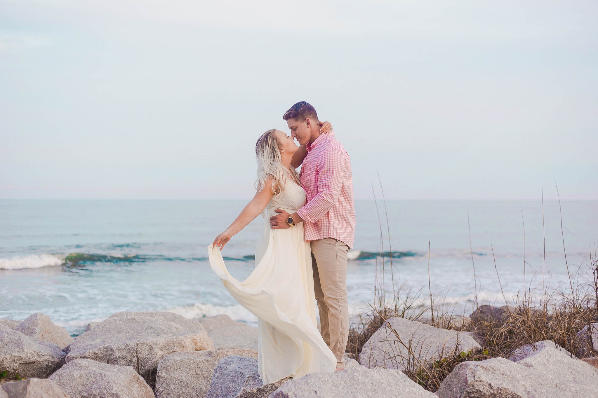 Engagement Photography Session at the beach on top of rocks with a cotton candy sky in the background - couple is kissing - girl is wearing a white flowy maxi dress from lulus - Honolulu Oahu Hawaii Wedding Photographer - Johanna Dye