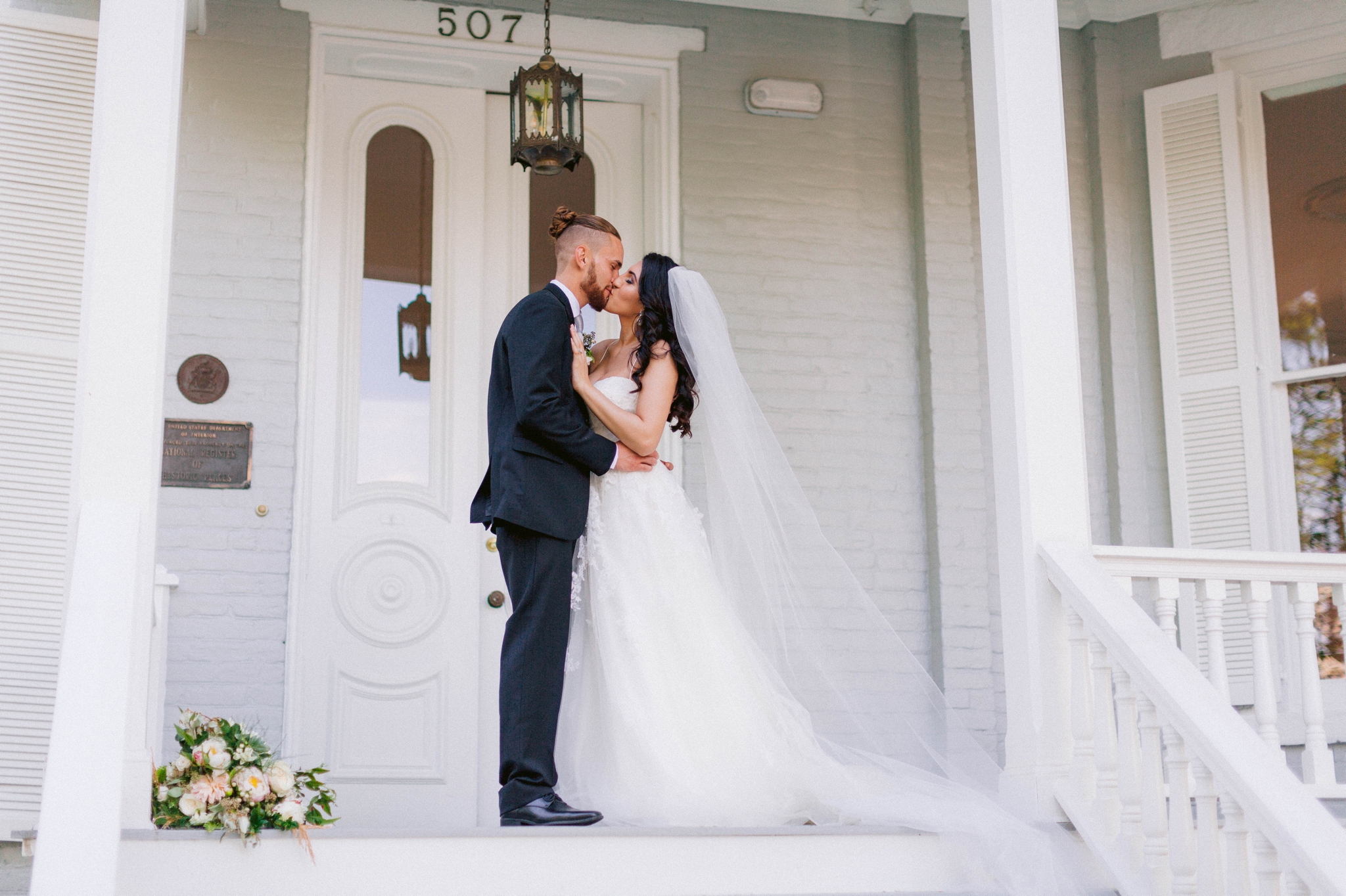  Couple Kissing - Wedding Portraits on the front porch of an all white luxury estate mansion - Bride is wearing a Aline Ballgown by Cherish by Southern Bride with a long cathedral veil - Groom is wearing a black suit by Generation Tux and has a man b