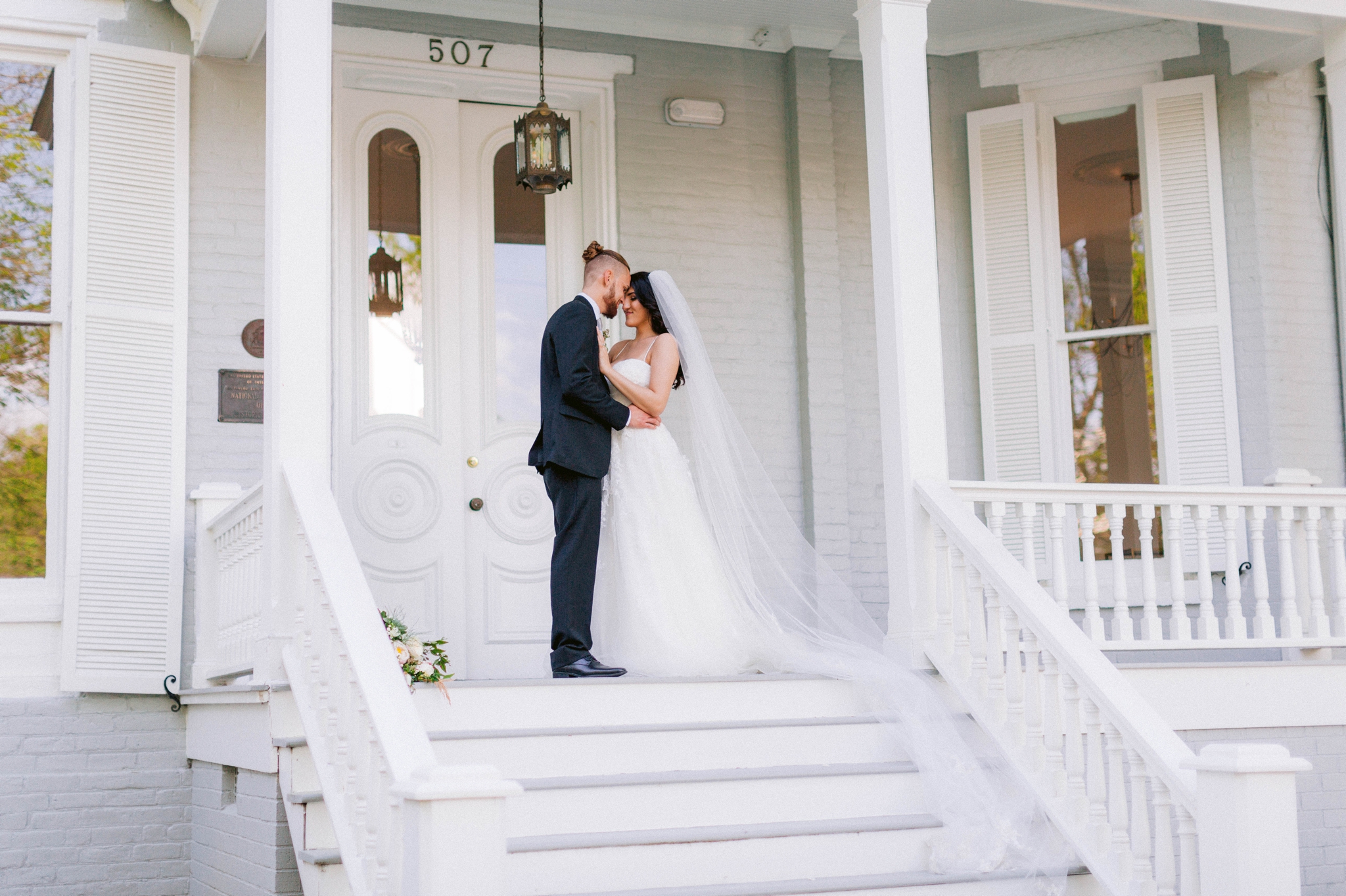  Couple Kissing - Wedding Portraits on the front porch of an all white luxury estate mansion - Bride is wearing a Aline Ballgown by Cherish by Southern Bride with a long cathedral veil - Groom is wearing a black suit by Generation Tux and has a man b