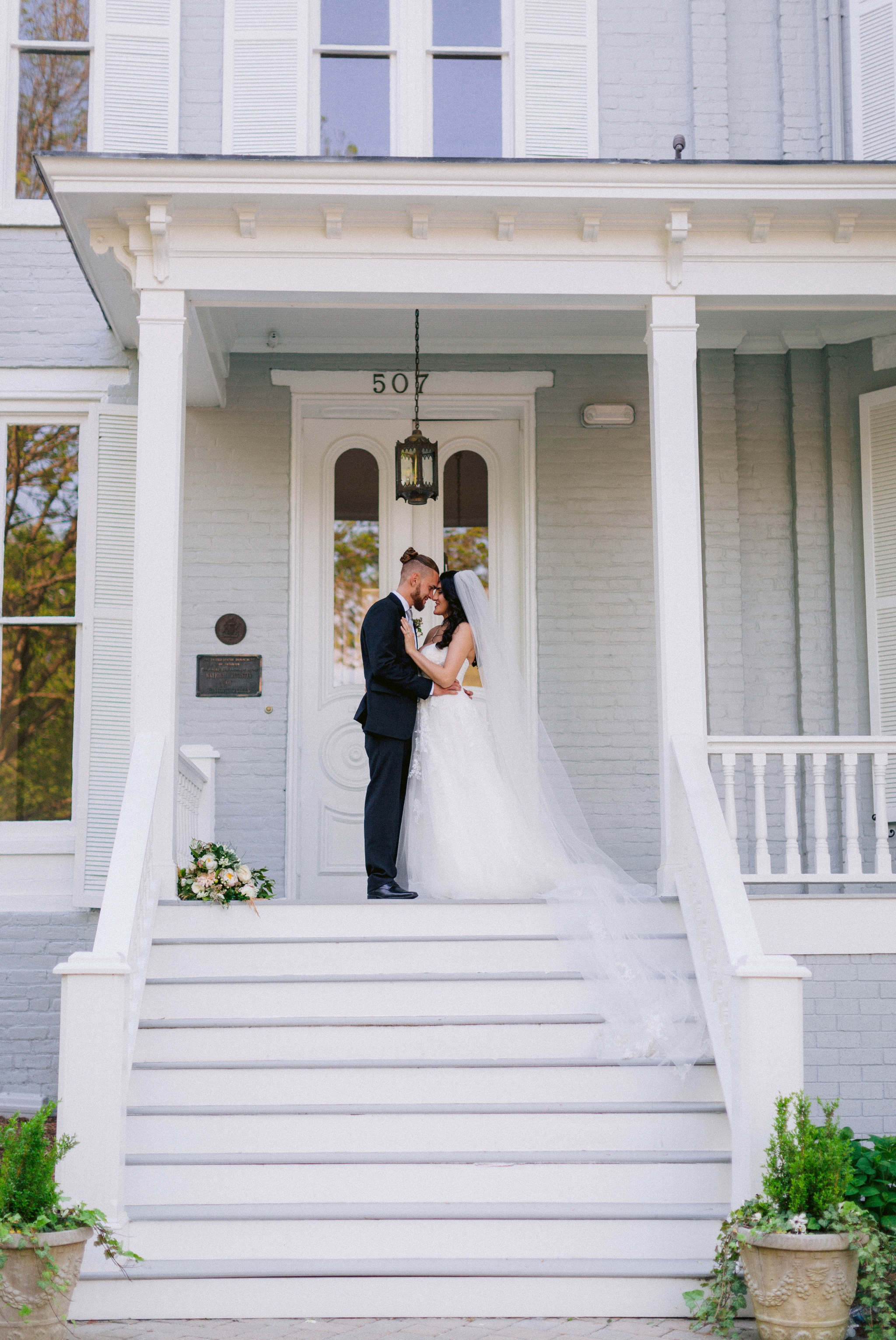  Couple Kissing - Wedding Portraits on the front porch of an all white luxury estate mansion - Bride is wearing a Aline Ballgown by Cherish by Southern Bride with a long cathedral veil - Groom is wearing a black suit by Generation Tux and has a man b