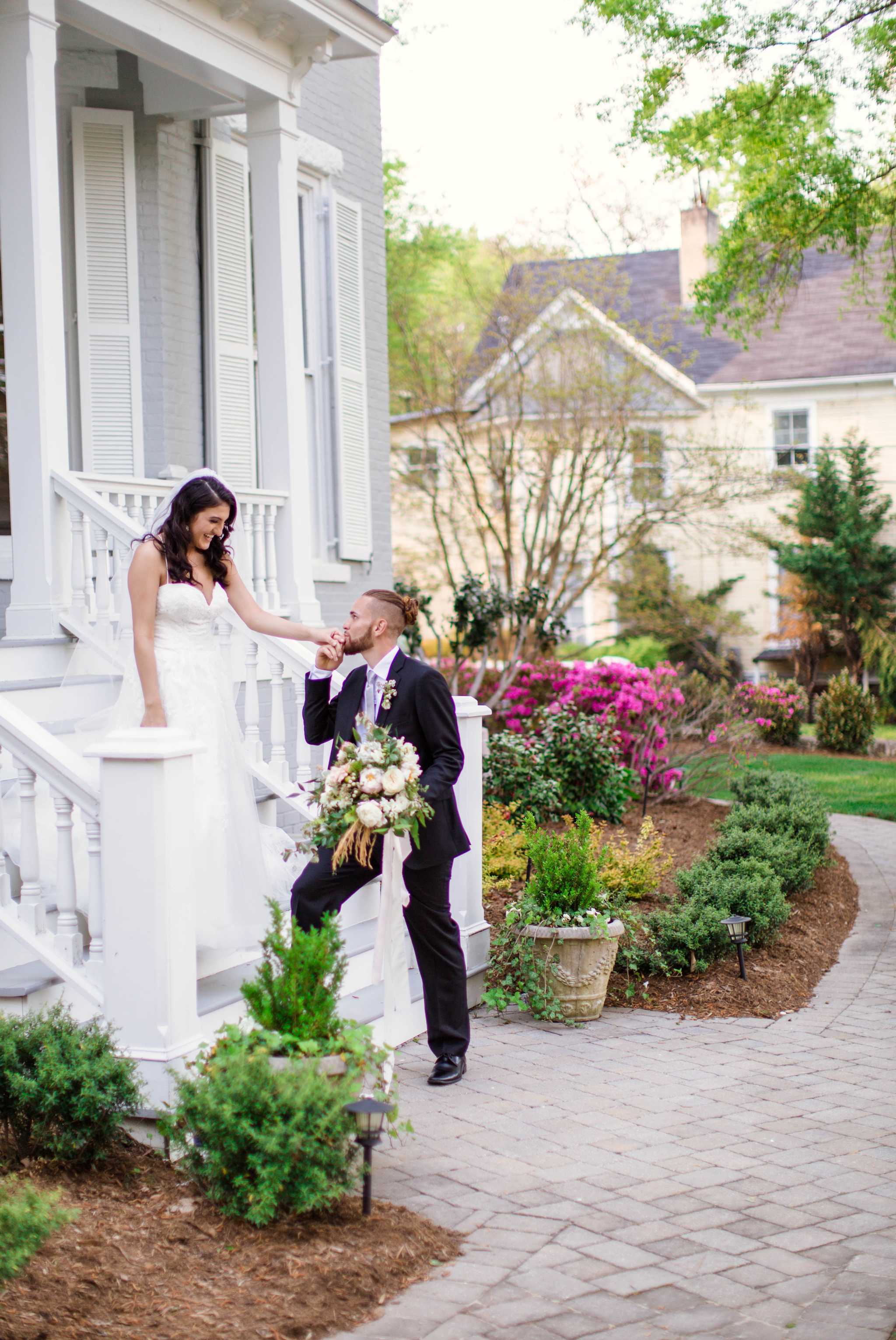  Groom kissing his brides hand  - Wedding Portraits on the front porch of an all white luxury estate mansion - Bride is wearing a Aline Ballgown by Cherish by Southern Bride with a long cathedral veil - Groom is wearing a black suit by Generation Tux