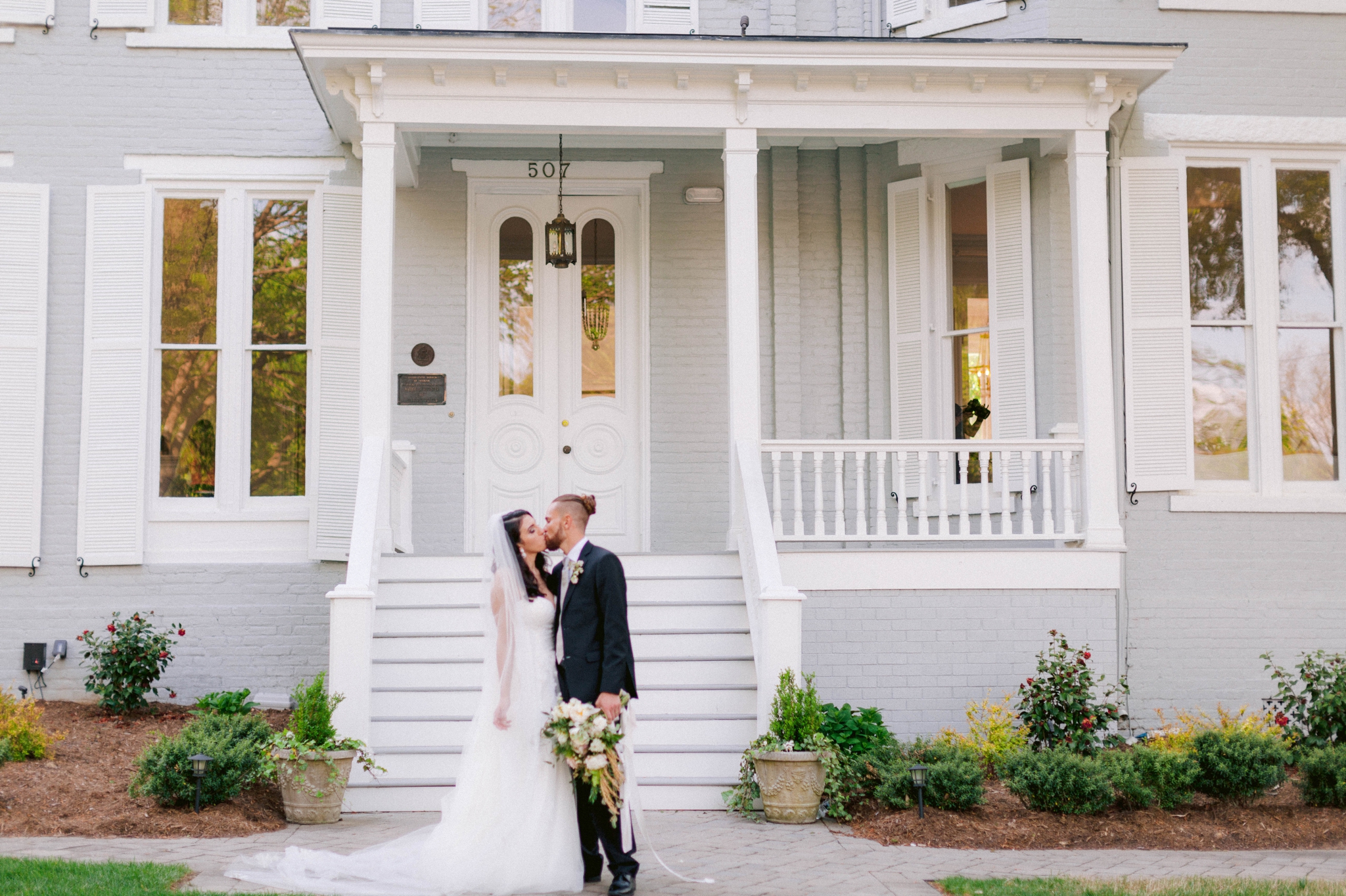  Couple Kissing in front of the mansion - Wedding Portraits on the front porch of an all white luxury estate  - Bride is wearing a Aline Ballgown by Cherish by Southern Bride with a long cathedral veil - Groom is wearing a black suit by Generation Tu